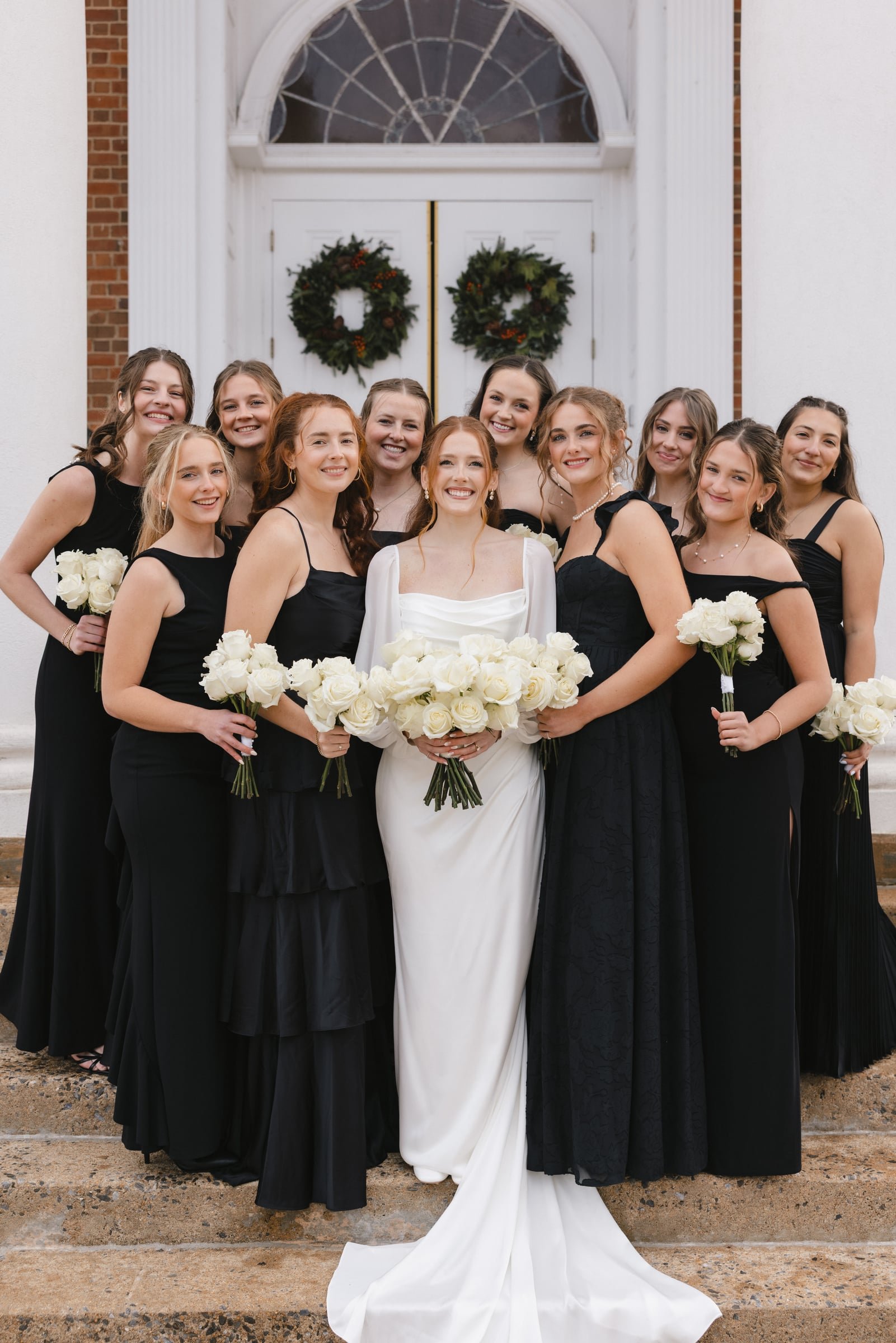 Group of women in black dresses with one bride in white, holding white roses, standing on steps outside a building with two wreaths on the door.