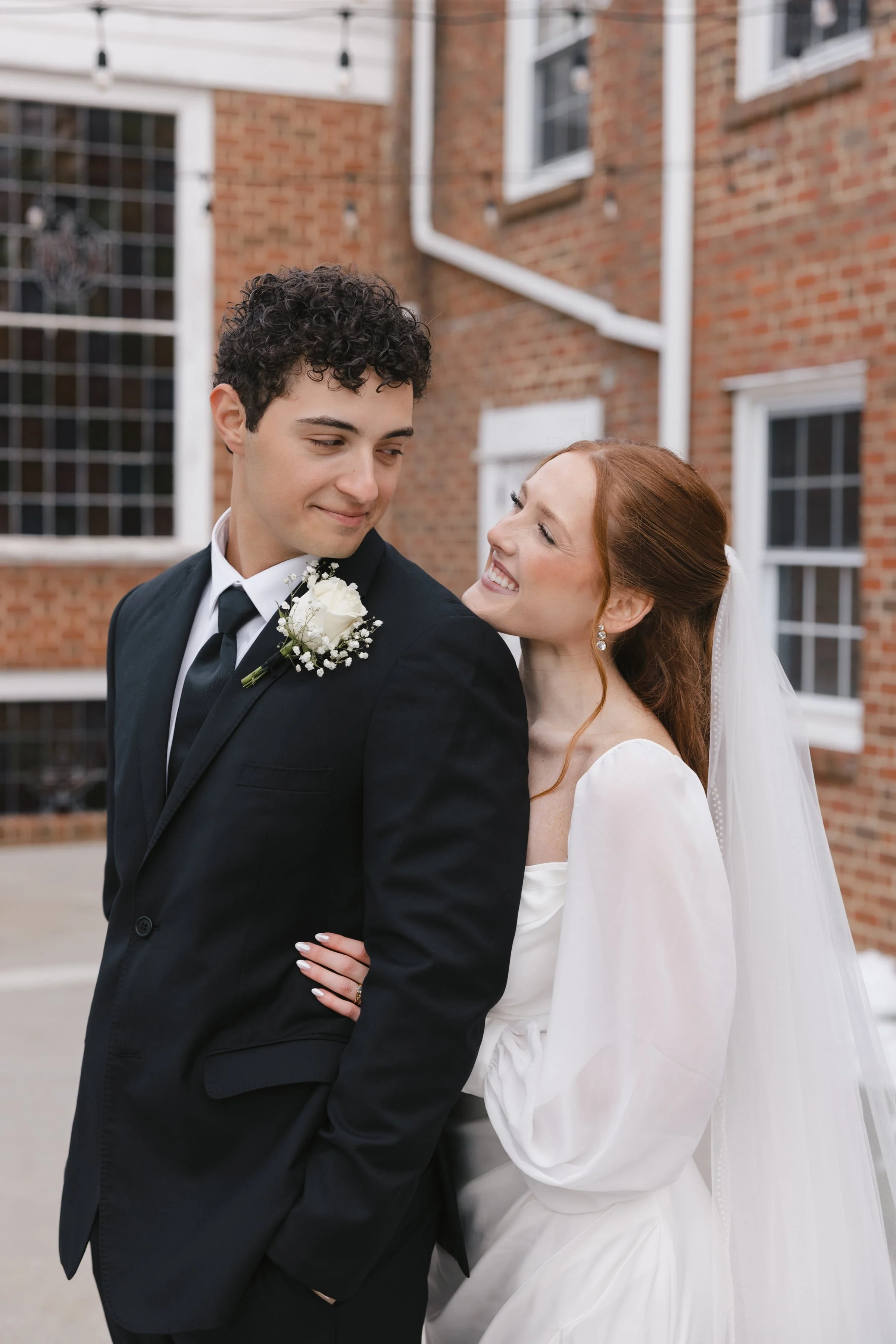 A happy couple dressed in wedding attire, standing close together outdoors in front of a brick building with large windows. The groom is wearing a black suit with a white shirt and black tie, and a white floral boutonniere. The bride has red hair sty
