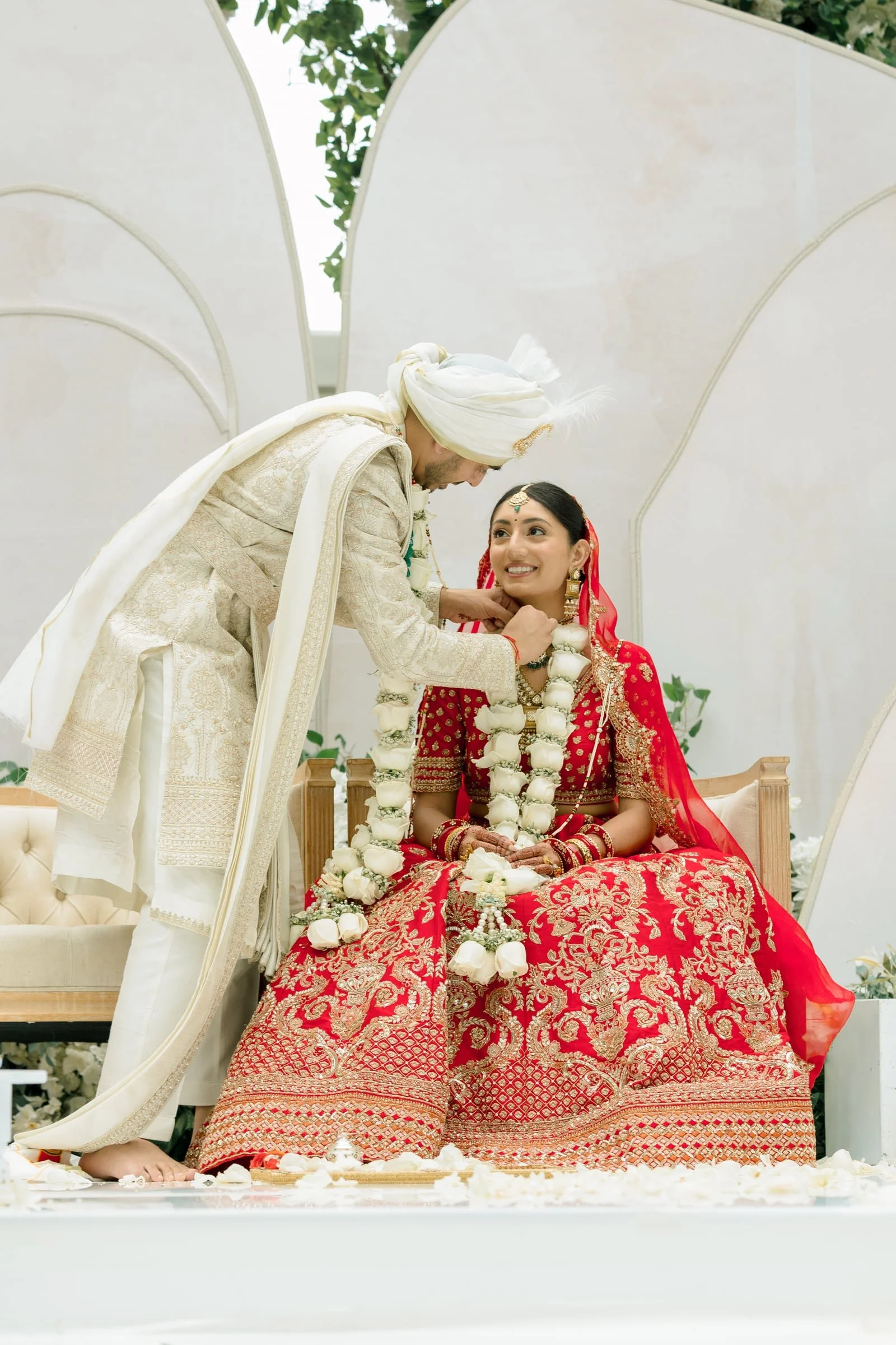 A traditional Indian wedding ceremony with a bride dressed in a red and gold embroidered outfit and a groom in a cream-colored sherwani and white turban, sitting during the ceremony surrounded by floral decorations and petals.