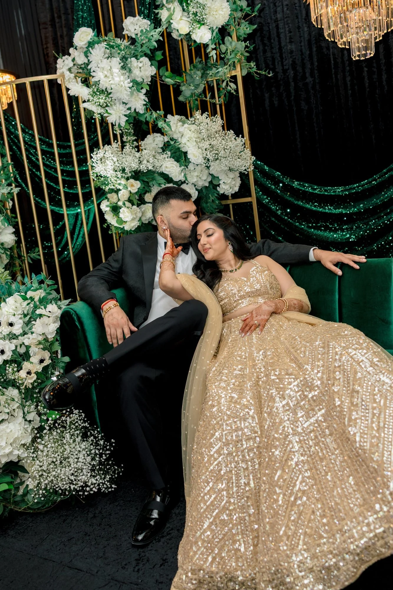 A young man and woman dressed in formal wedding attire, sitting closely on a green velvet couch, surrounded by white floral decorations, with the young man gently kissing the woman's forehead.