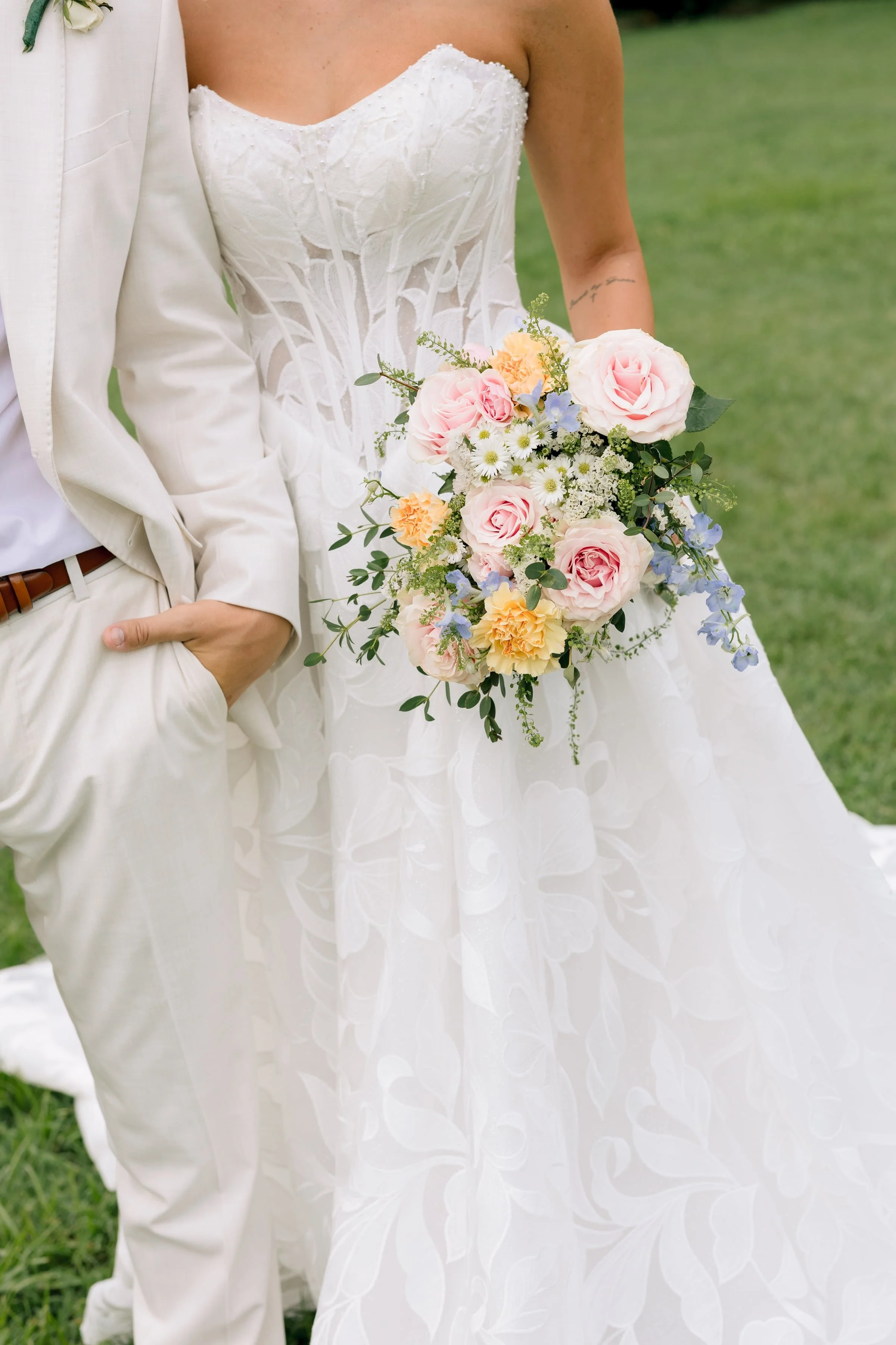Close-up of a bride holding a bouquet of pink roses, yellow carnations, white daisies, and greenery, wearing a white strapless wedding dress with textured floral patterns, standing outdoors on grass.