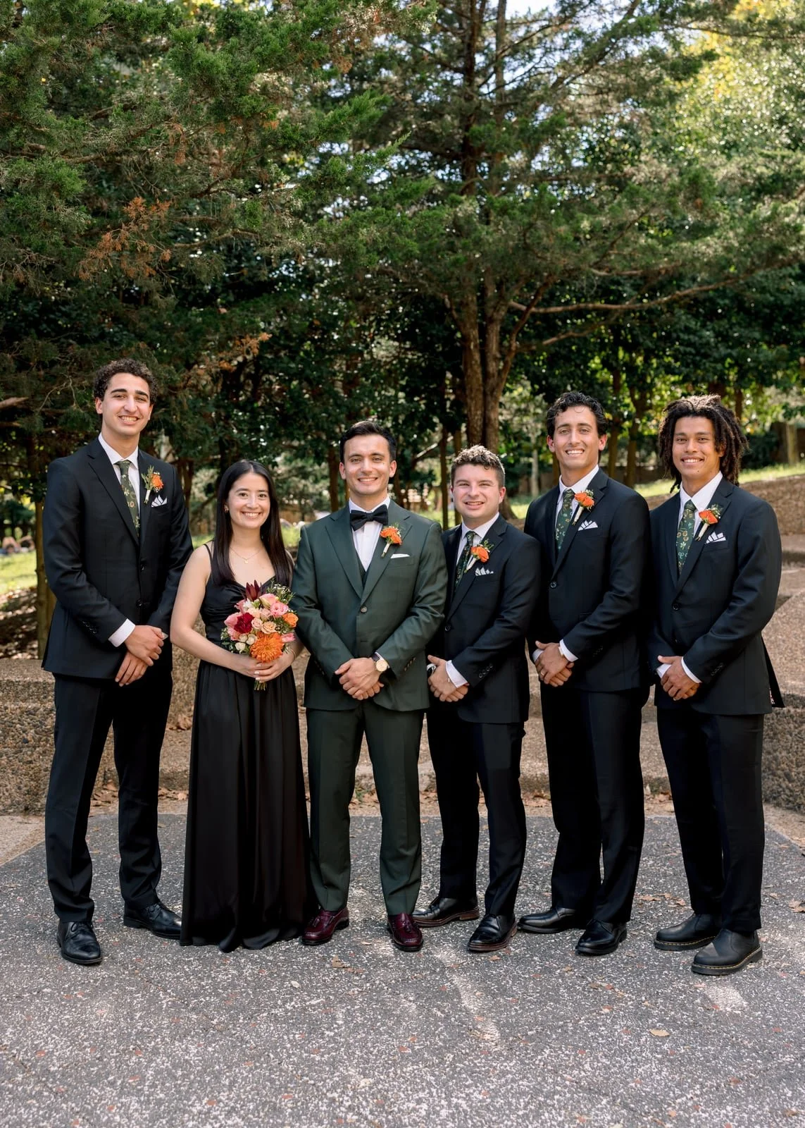 A group of six people dressed in formal attire, standing outdoors under trees, with one woman holding a bouquet of flowers.