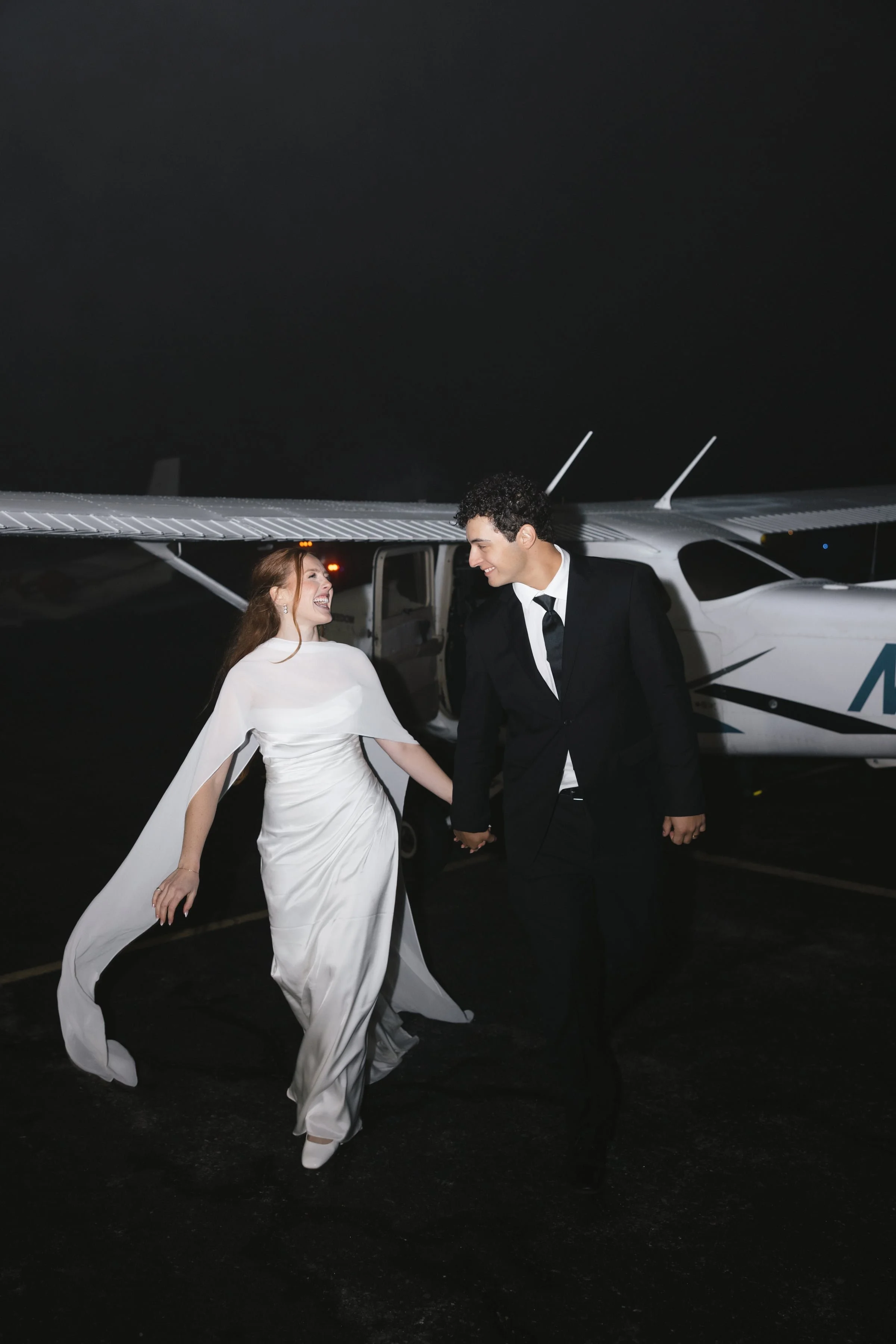 A newlywed couple, the bride in a white wedding gown and the groom in a black tuxedo, holding hands and smiling as they walk away from a small airplane on a dark night.