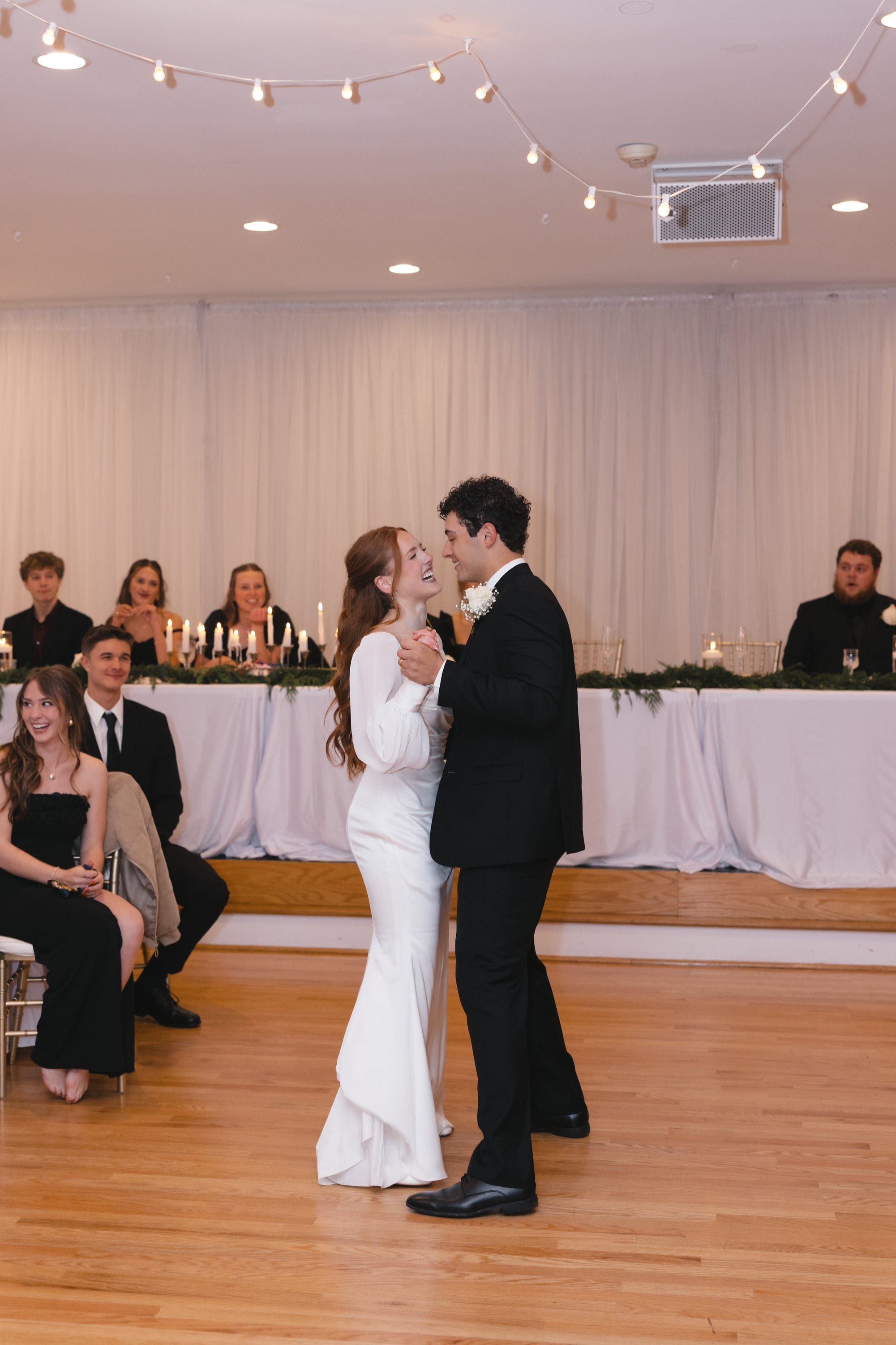 A bride and groom share a dance during their wedding reception, with guests watching and smiling in the background.