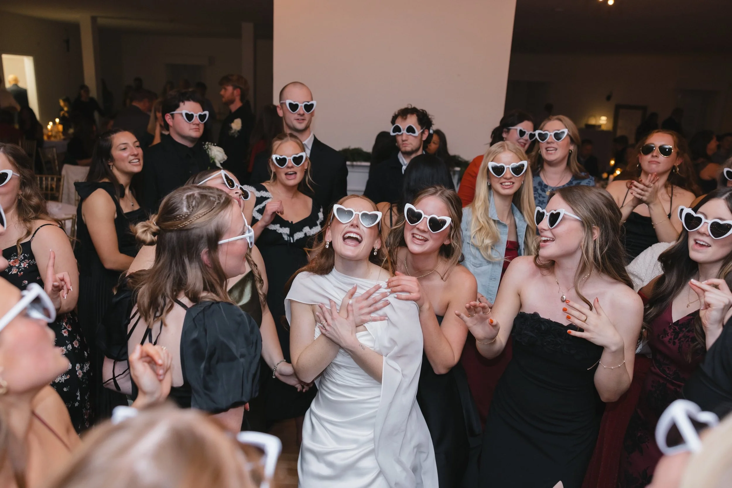 Group of people at an indoor event wearing white heart-shaped sunglasses, smiling and having fun.
