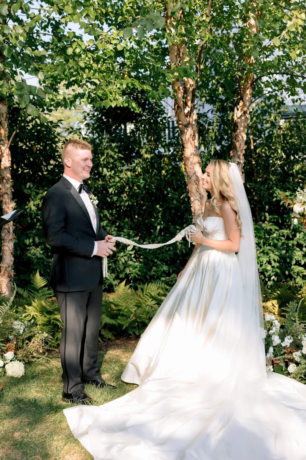 A bride and groom exchanging vows outdoors, holding a rope together, surrounded by green trees and plants.