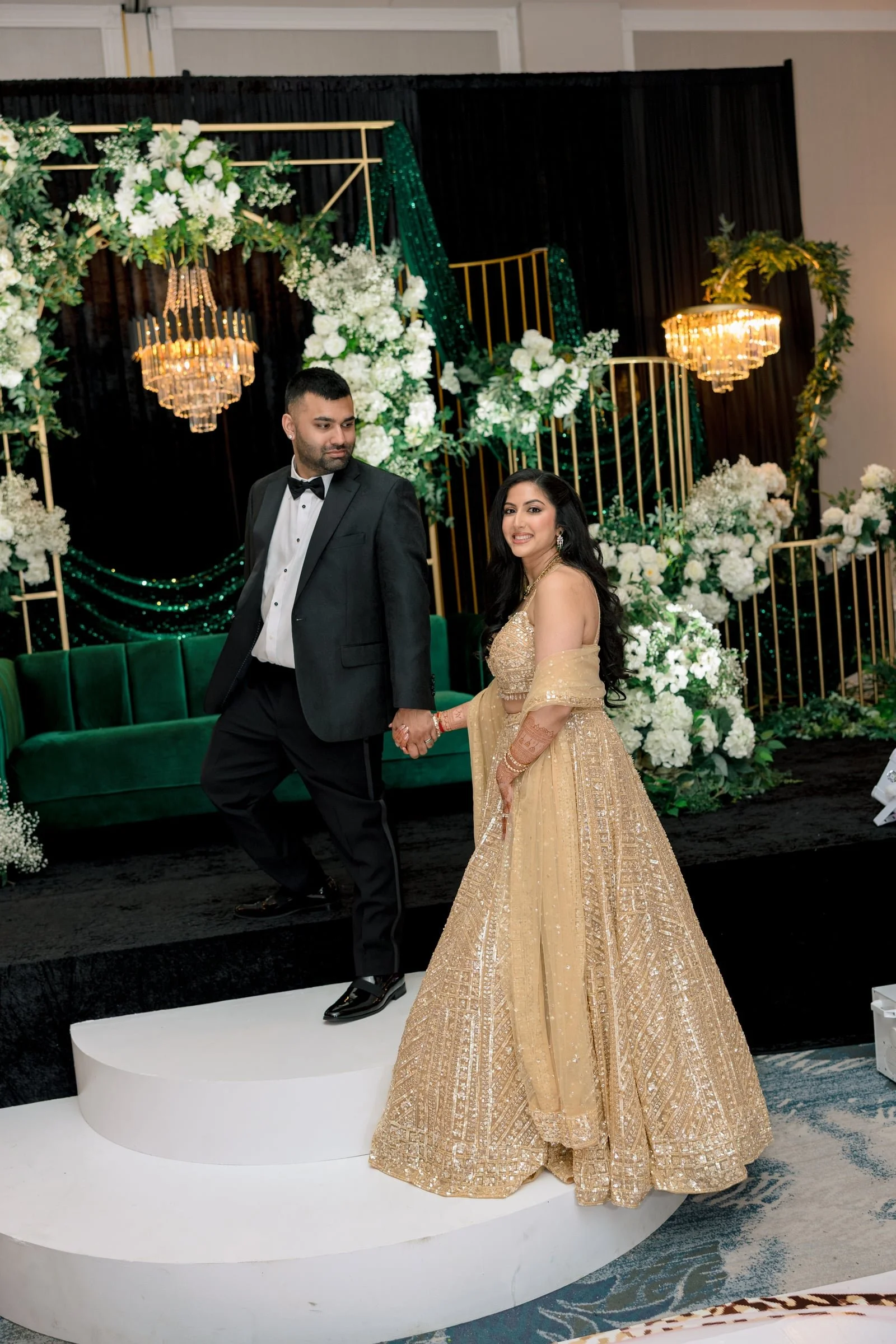 A couple at their formal wedding celebration, holding hands as they stand on a white podium with a black and green set decorated with white flowers and gold accents in the background. The woman is wearing a gold traditional dress, and the man is in a