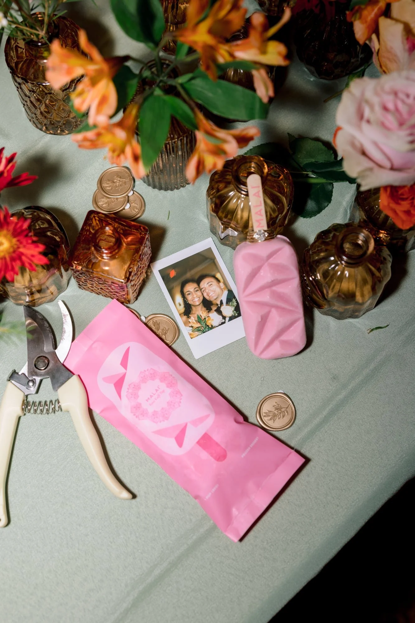 A table decorated with pink and orange flowers in brown vases, pink and gold wedding decor, a romantic pink soap bar, a polaroid photo of a smiling couple, and rose gold wedding coins.