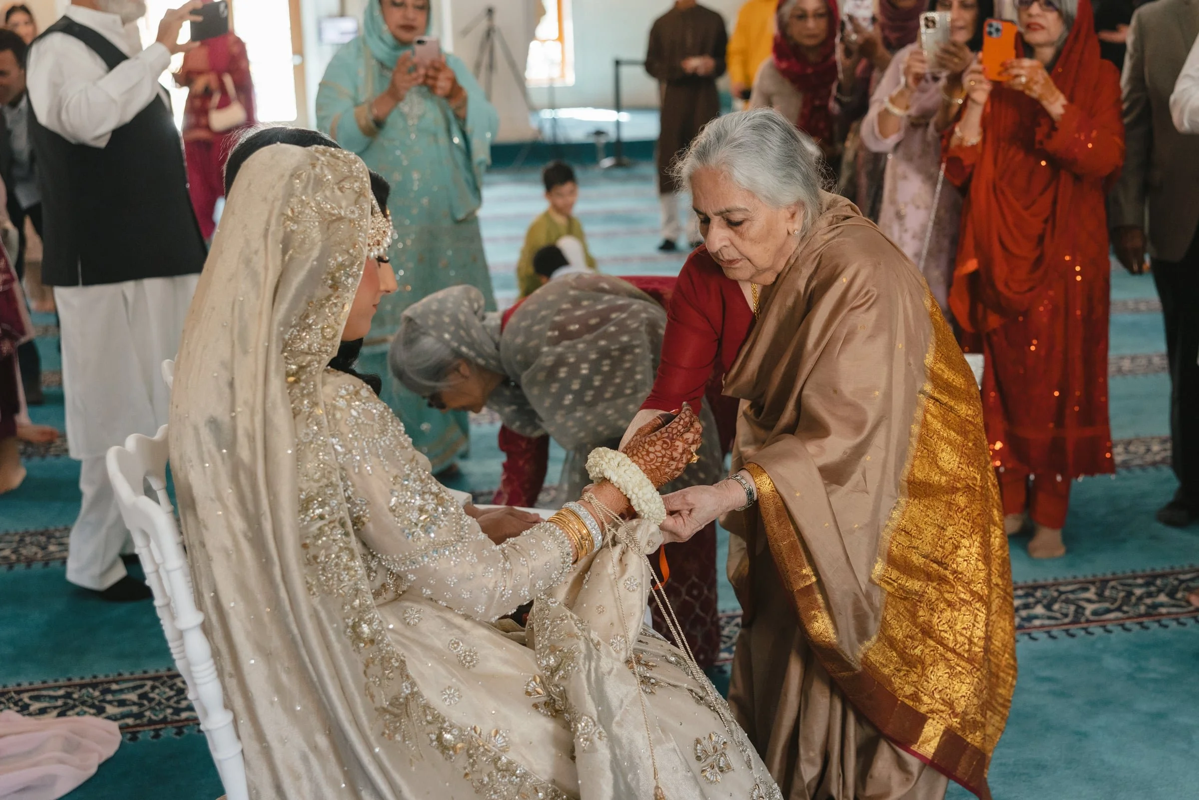 An elderly woman dressed in traditional clothing is holding the hand of a bride in an elegant gown, during a wedding ceremony. Several guests are standing around, some taking photos, in a decorated indoor venue.