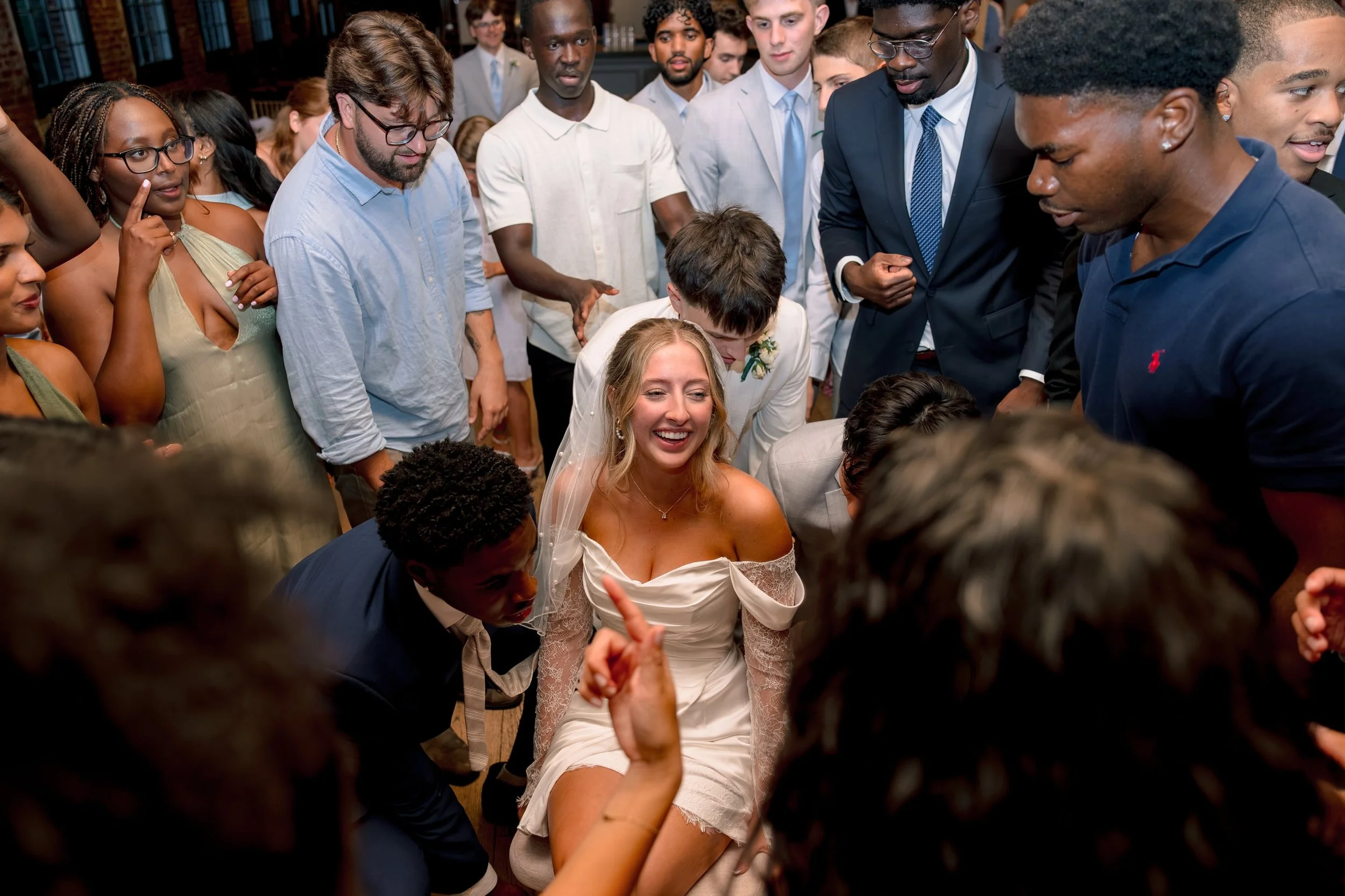 A group of diverse people surrounding and celebrating a woman in a white wedding dress, who is smiling and sitting on the floor, during a lively wedding reception or celebration.