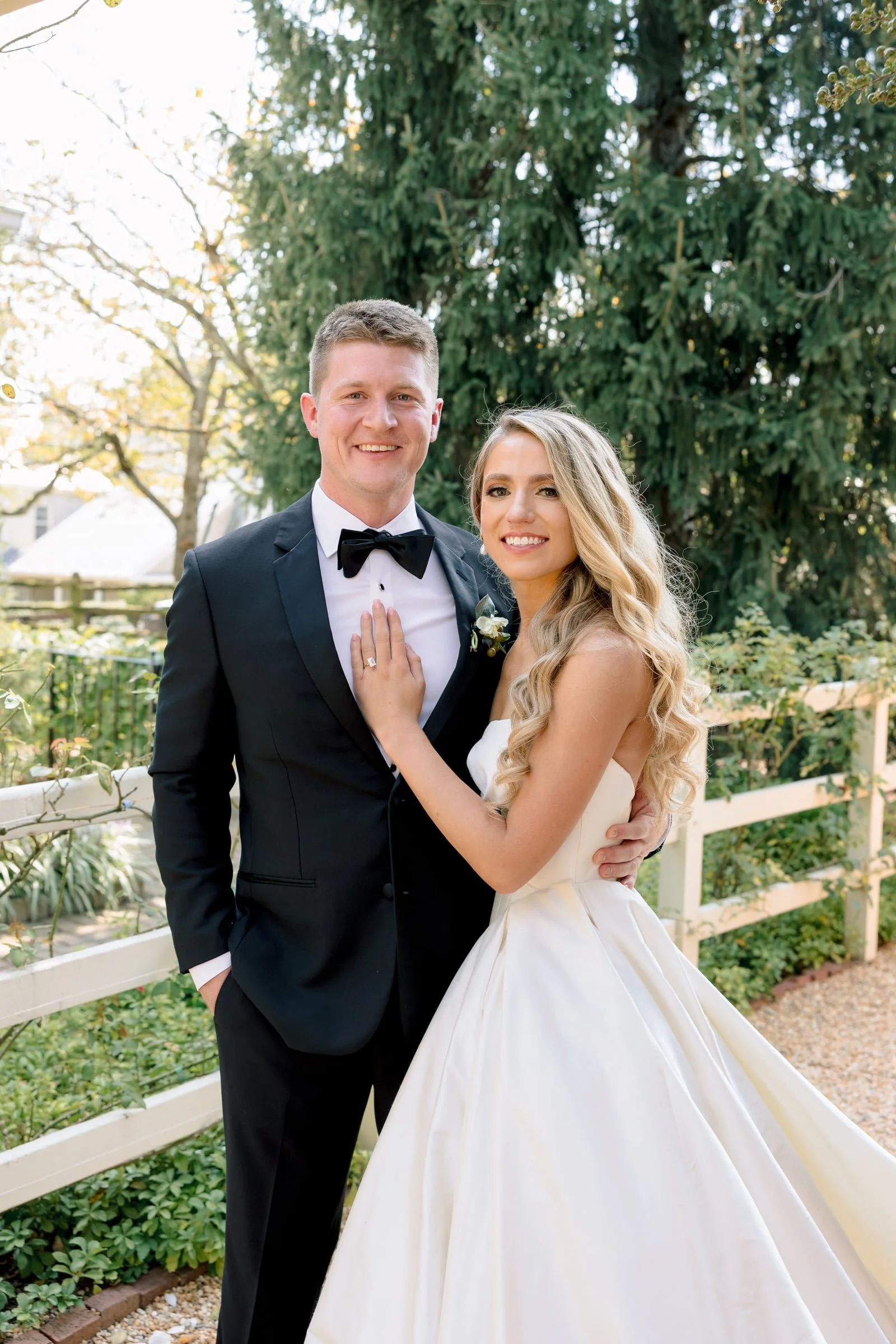 A smiling man and woman dressed in wedding attire standing outdoors near a white fence and green foliage.