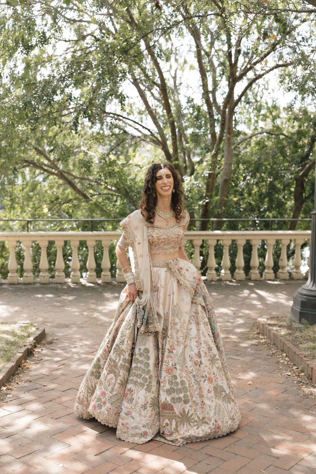 A woman dressed in traditional Indian wedding attire, standing outdoors on a brick pathway with green trees and a decorative stone railing in the background. She is smiling and wearing an ornate cream-colored lehenga with intricate embroidery, jewelr