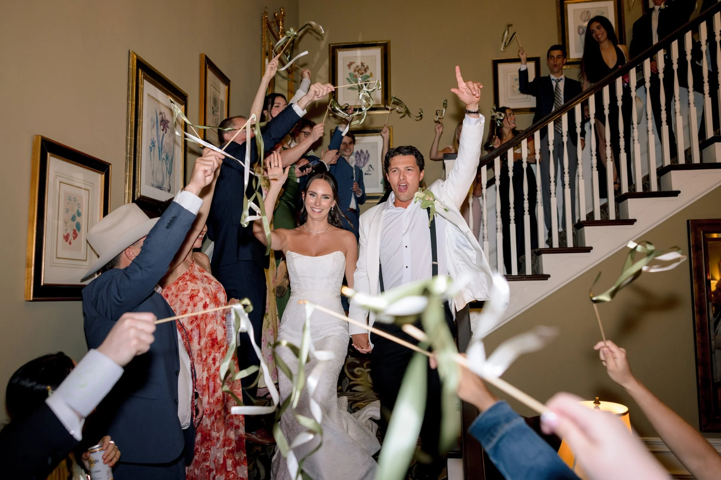 A bride and groom celebrating with wedding guests during a party, holding hands and surrounded by guests hitting party blowers, on a staircase inside a decorated home.