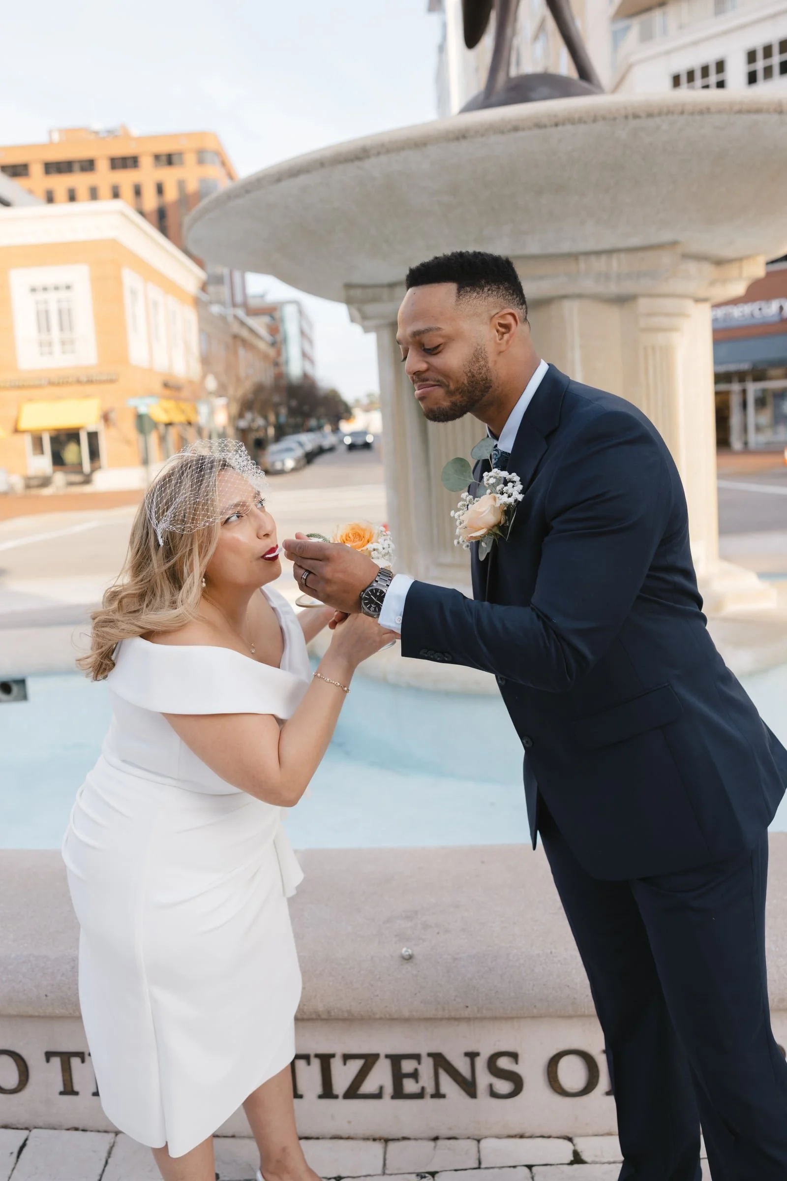 A man in a dark suit and a woman in a white dress, with a bird veil, sharing a moment in front of a fountain in a city street, with buildings in the background.