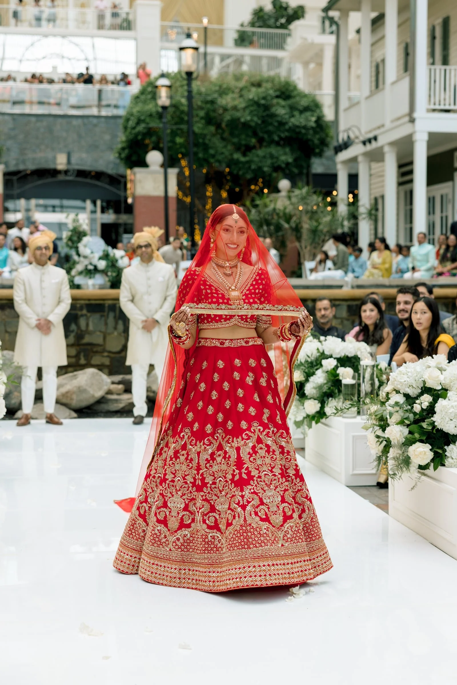 A bride in a red and gold traditional Indian wedding dress, holding a gold plate, standing during her wedding ceremony with guests and a decorated background behind her.