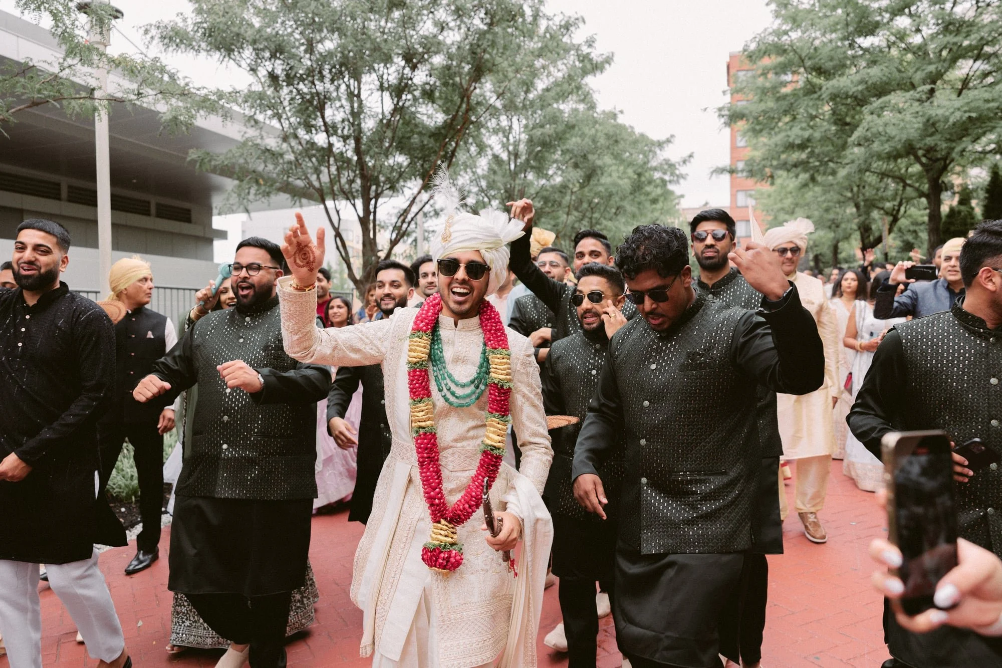 A group of people celebrating at an outdoor event, with a man in traditional Indian attire wearing sunglasses and a turban, decorated with flower garlands, dancing and smiling.