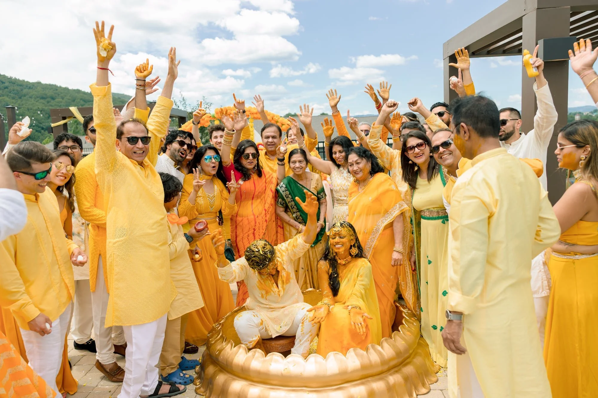 Celebration of an Indian wedding with guests dressed in yellow and orange clothes, some with colorful accessories, gathered around the bride and groom who are seated and covered in yellow powder, indicating a traditional pre-wedding ritual outdoors in sunny weather.