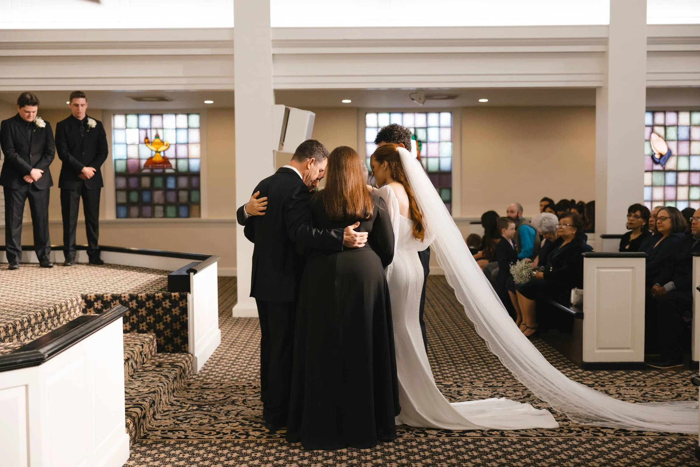 A bride and groom kneeling in prayer with two women in an indoor church setting during a wedding ceremony, with guests seated in the background and stained glass windows on the wall.