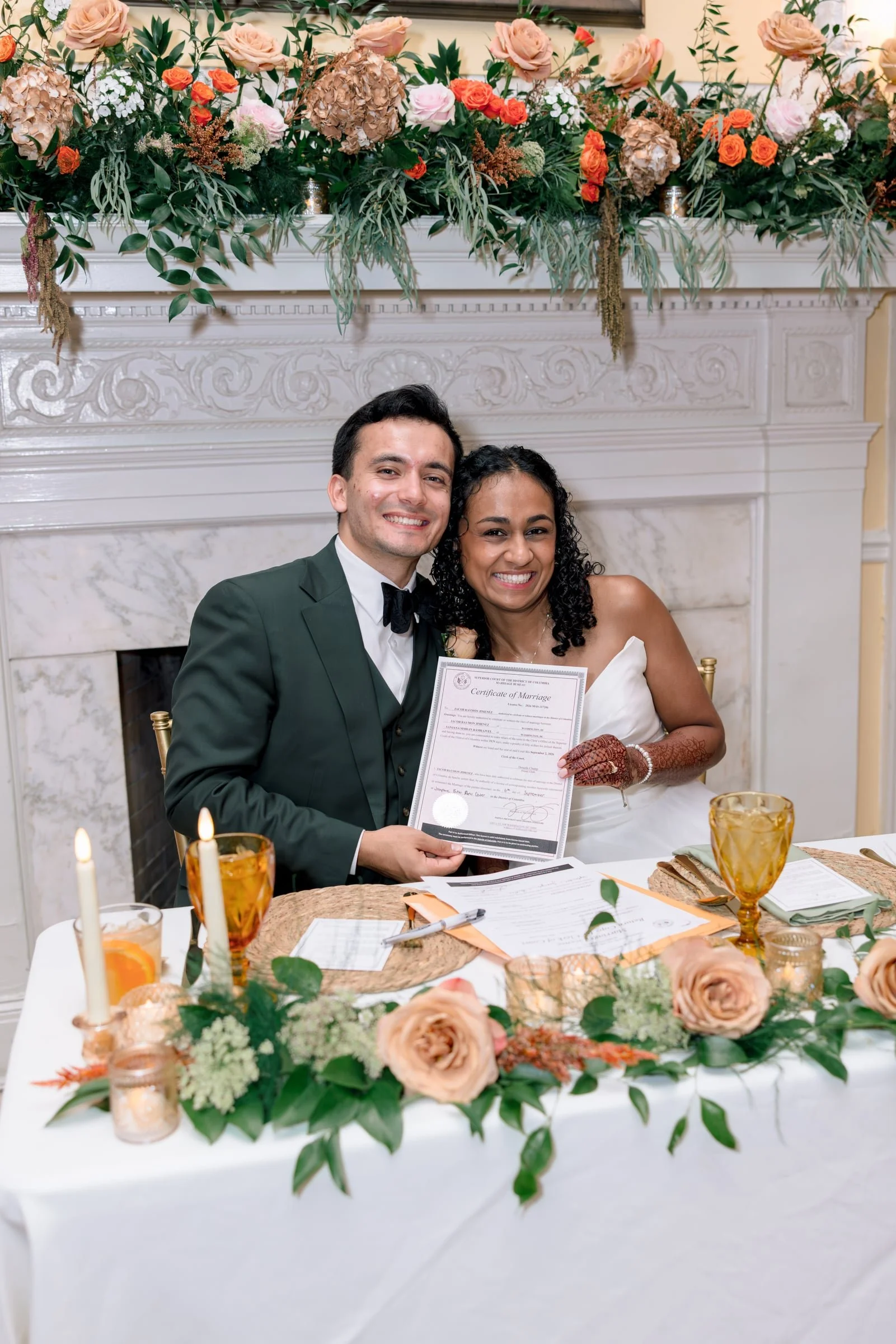 A newly married couple is smiling and holding a marriage certificate at a wedding reception. The man is wearing a dark green tuxedo with a black bow tie, and the woman is dressed in a white wedding gown with henna on her hands. They are sitting at a 