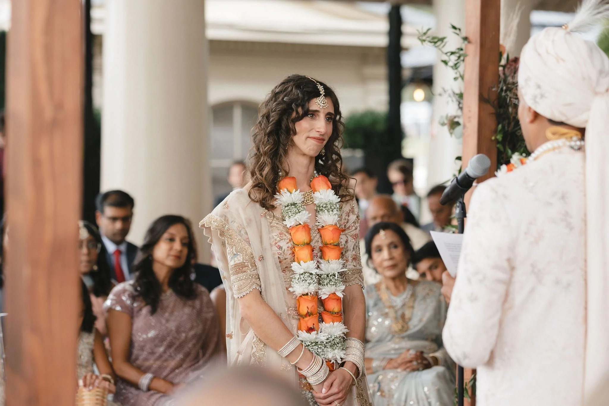 A woman dressed in traditional Indian attire with a garland of flowers around her neck, standing during a wedding ceremony as she looks at another woman who appears to be officiating. Several guests are seated in the background.