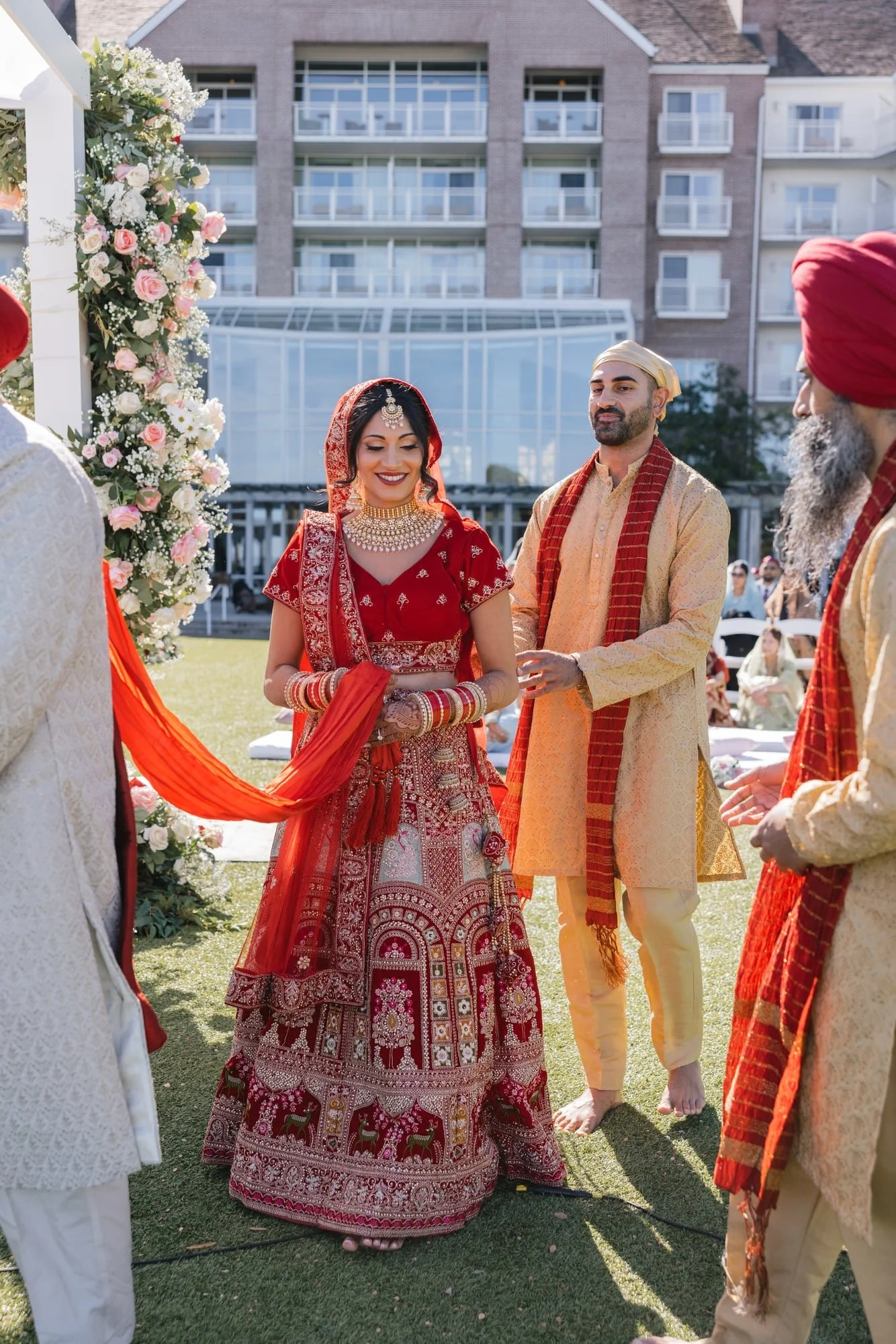 Indian wedding ceremony with a bride in red traditional attire and gold jewelry, surrounded by men in traditional Indian clothing, outdoors under a floral arch with a building in the background.