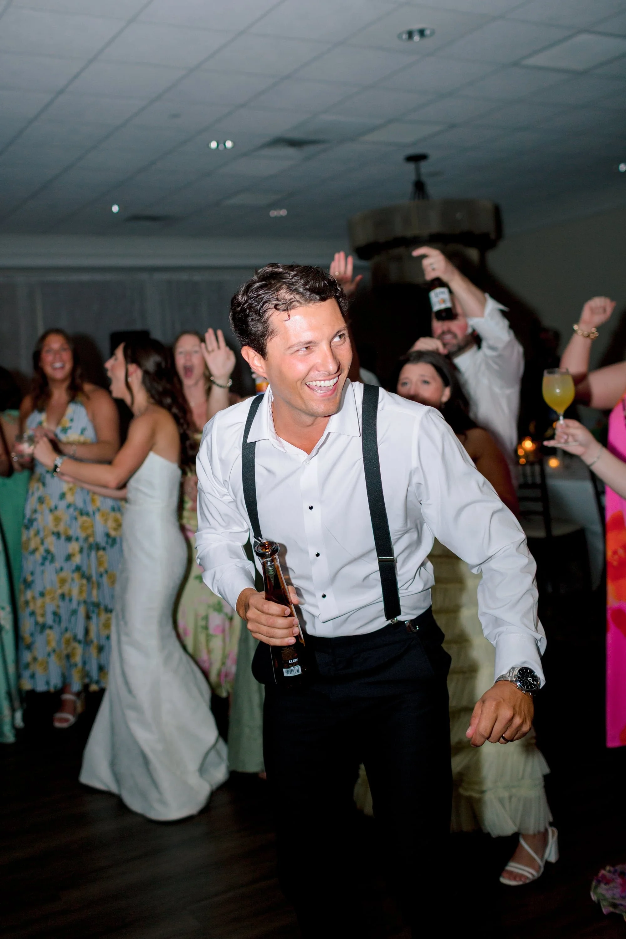A man in formal attire dancing at a celebration with a group of women and men in the background, some holding drinks and raising their hands, in a decorated indoor venue.
