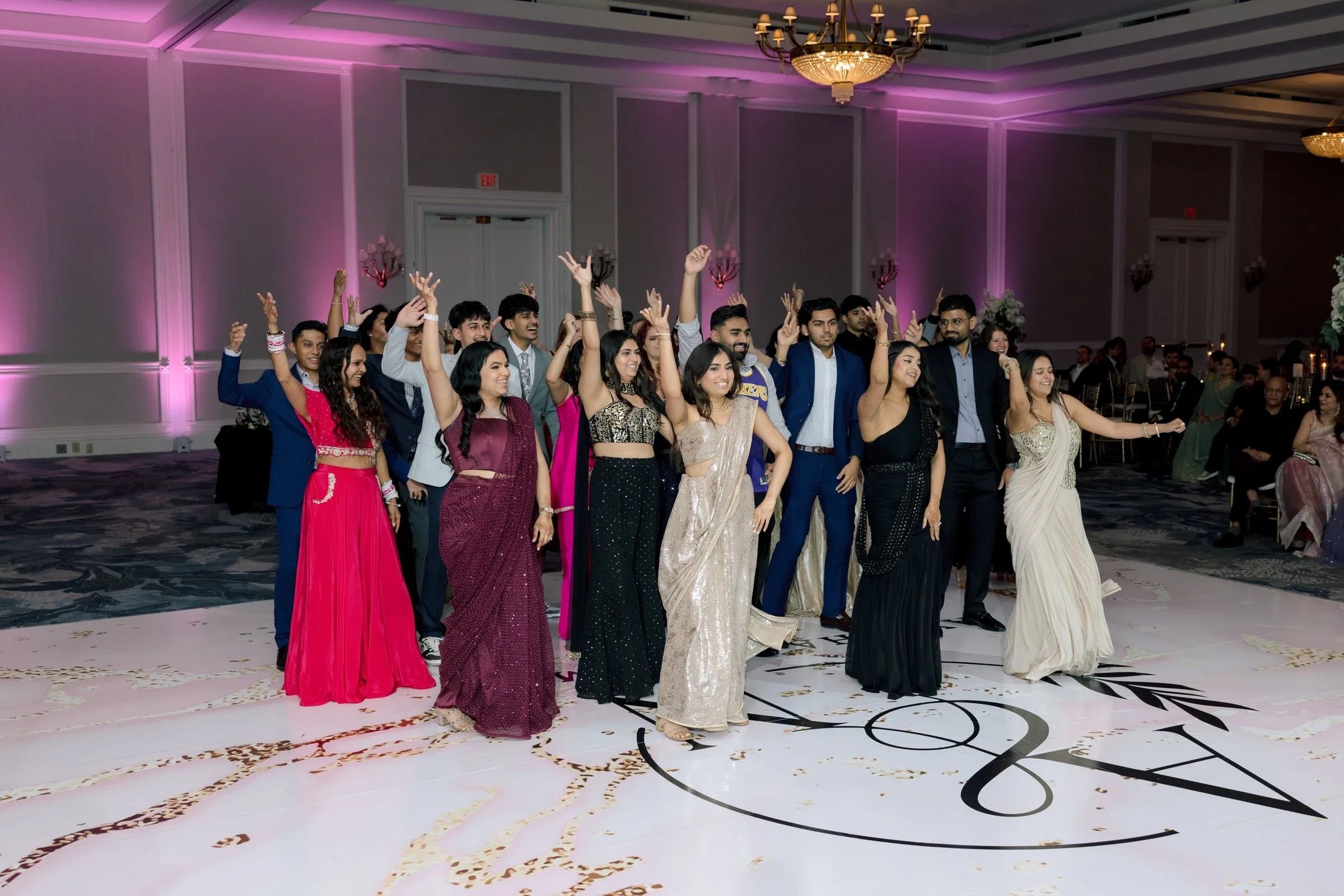 Group of people dancing and celebrating at a wedding reception in a decorated banquet hall with purple lighting and chandeliers.