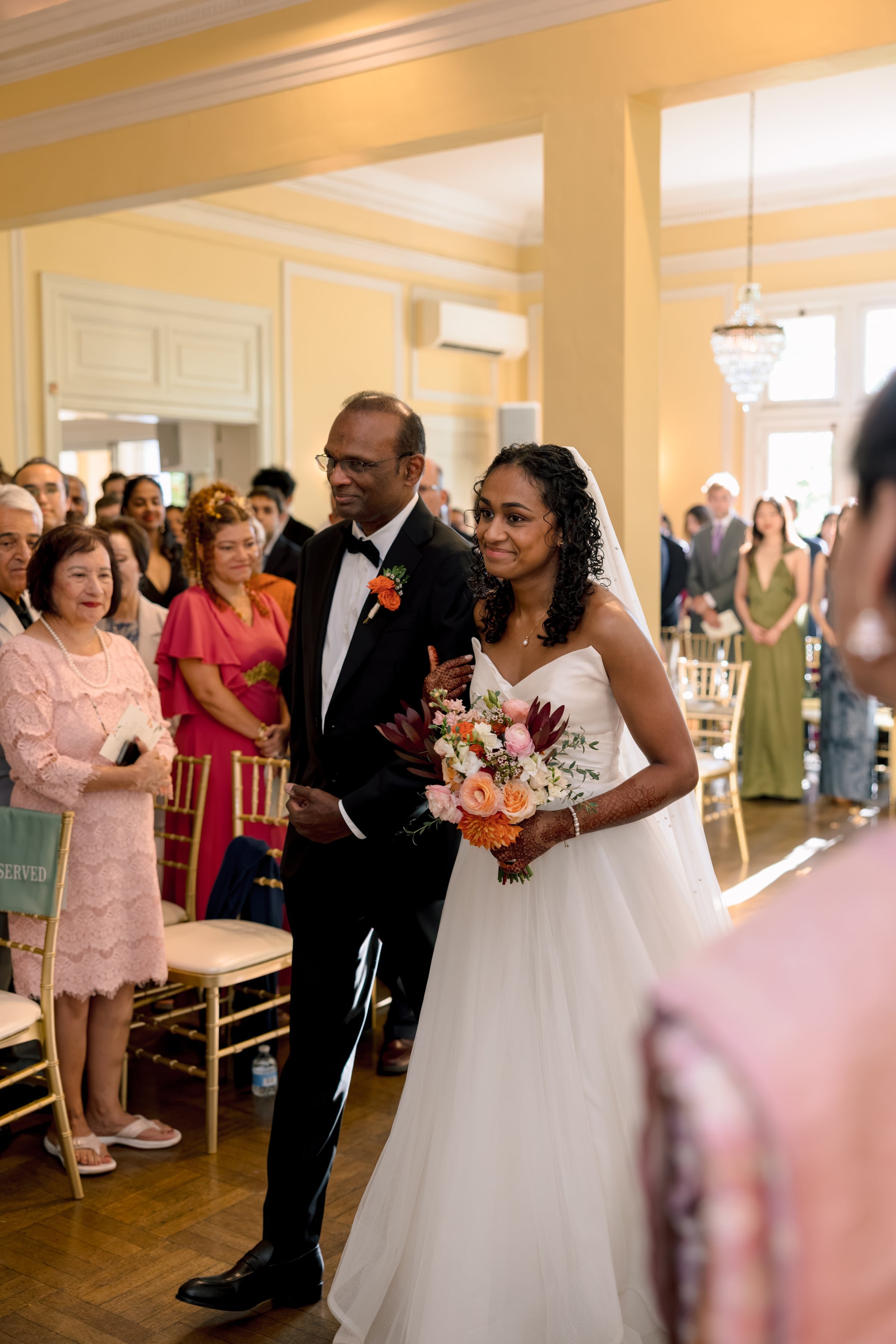A bride walking down the aisle with her father at a wedding ceremony, surrounded by seated guests in a decorated indoor venue.