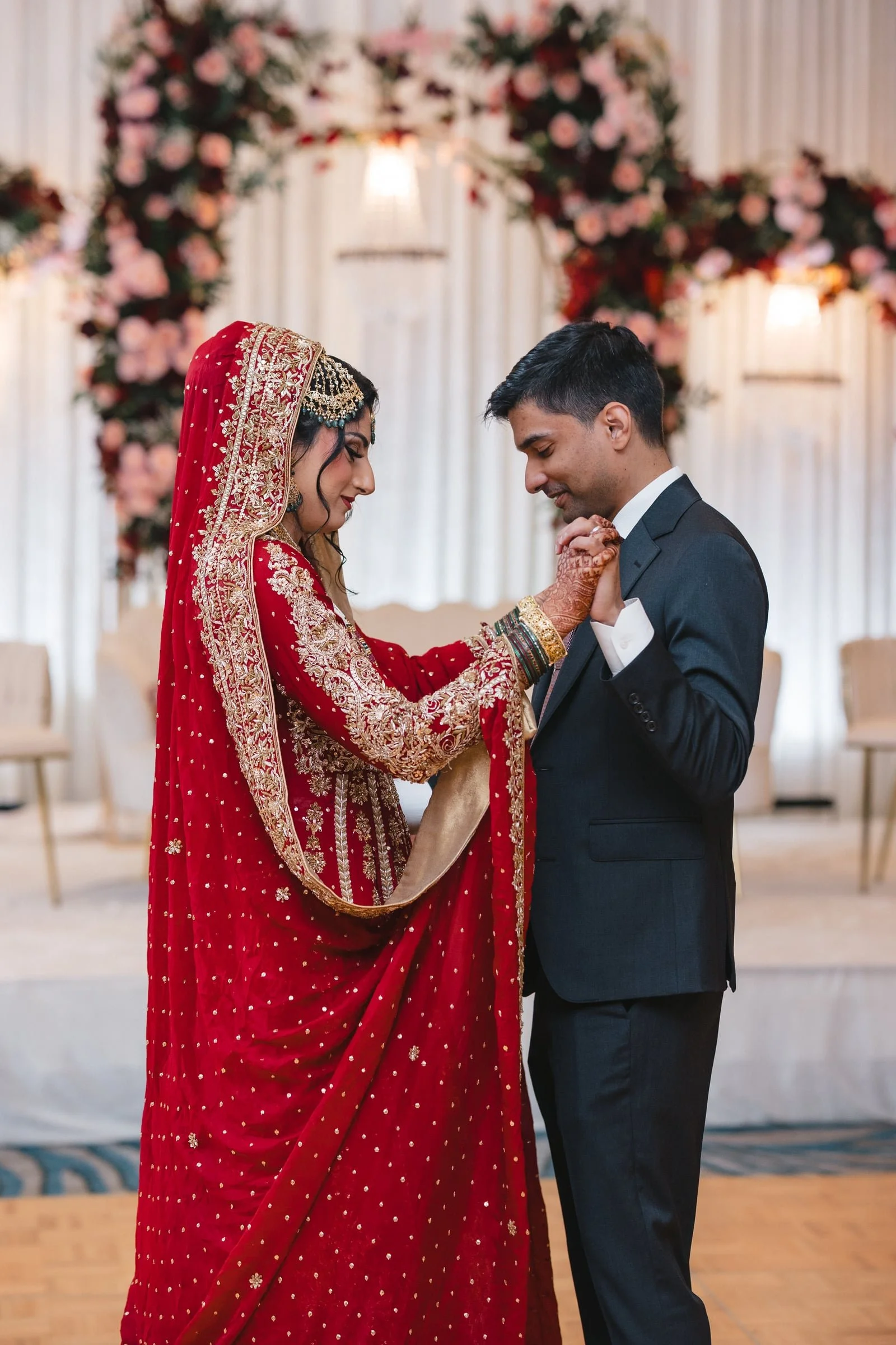 A bride and groom share a dance at their wedding reception. The bride wears a red traditional dress with gold embroidery and jewelry, and the groom is in a black suit.