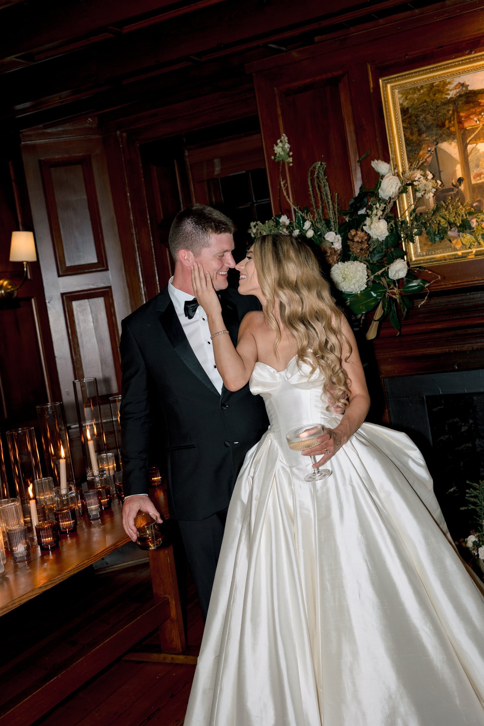 A bride and groom sharing a moment in a dimly lit, wood-paneled room with candles on a side table and a large floral arrangement behind them.