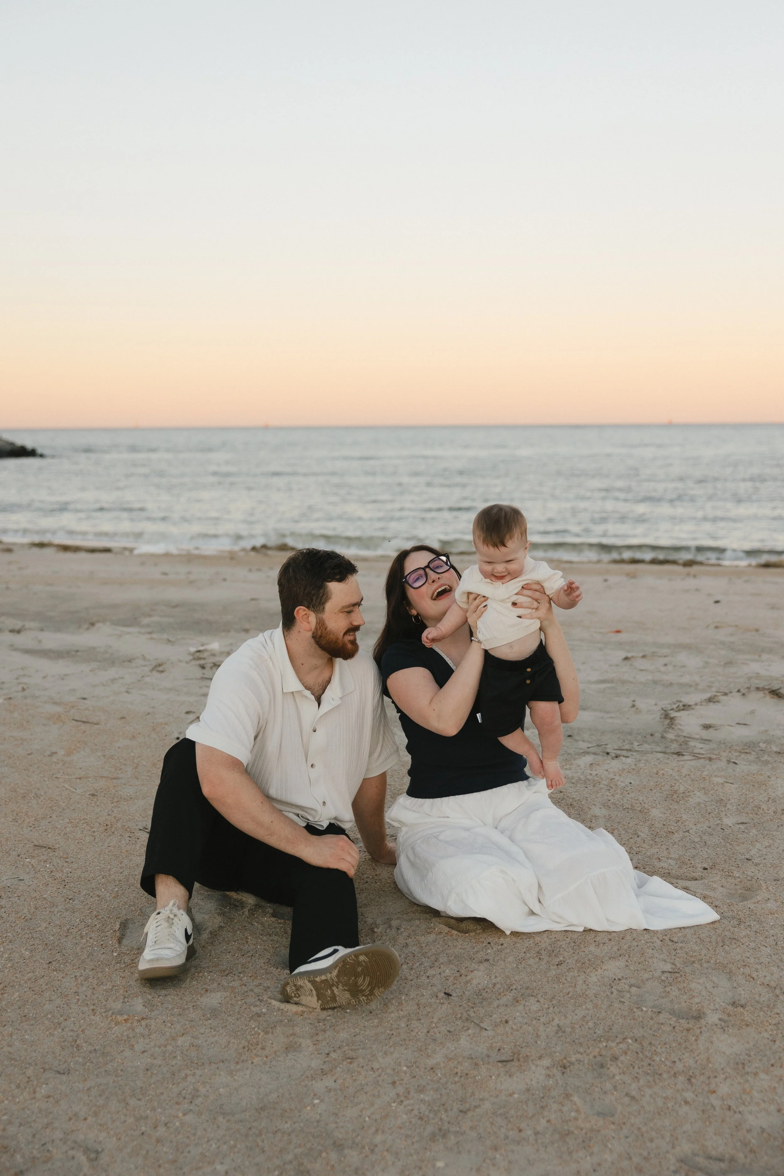 A family of three, including a father, a mother, and a baby, enjoying a day at the beach during sunset. The father is kneeling on the sand, smiling, while the mother is sitting and holding the baby, both smiling and laughing.