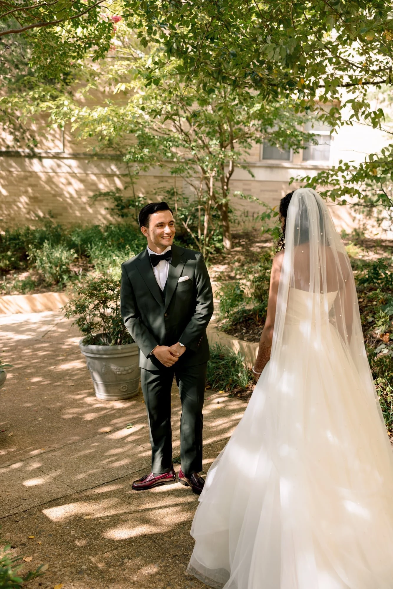 A groom in a black tuxedo and a bride in a white wedding gown and veil standing outdoors under trees, smiling at each other during their wedding.