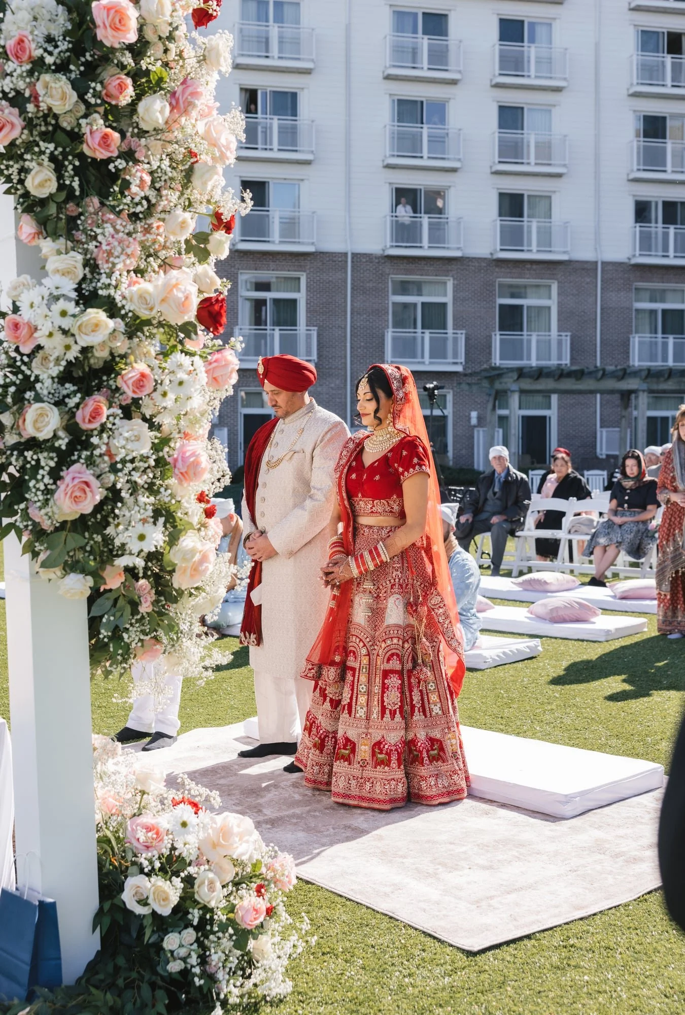 A couple dressed in traditional Indian wedding attire standing during their wedding ceremony, with floral decorations and guests seated nearby on a grassy outdoor setting.