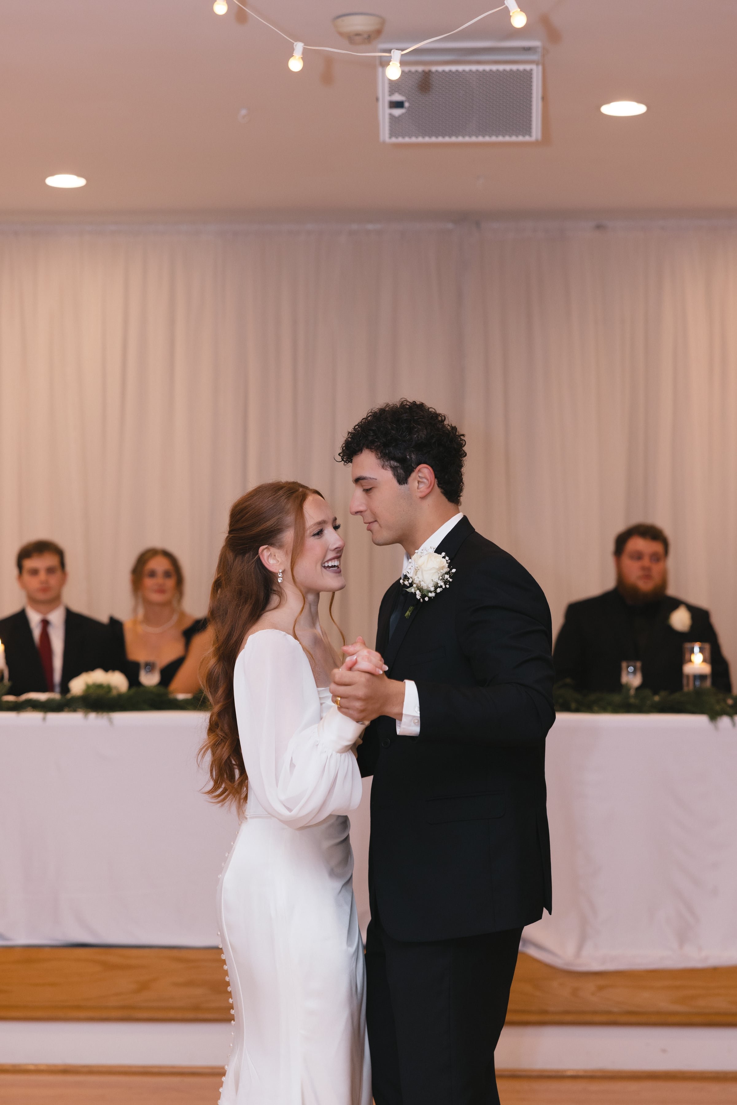 A couple is dancing at a wedding, with a bride in a white dress and a groom in a black tuxedo. They are smiling and looking at each other. In the background, there are five people dressed formally, including men in tuxedos and women in dresses, stand