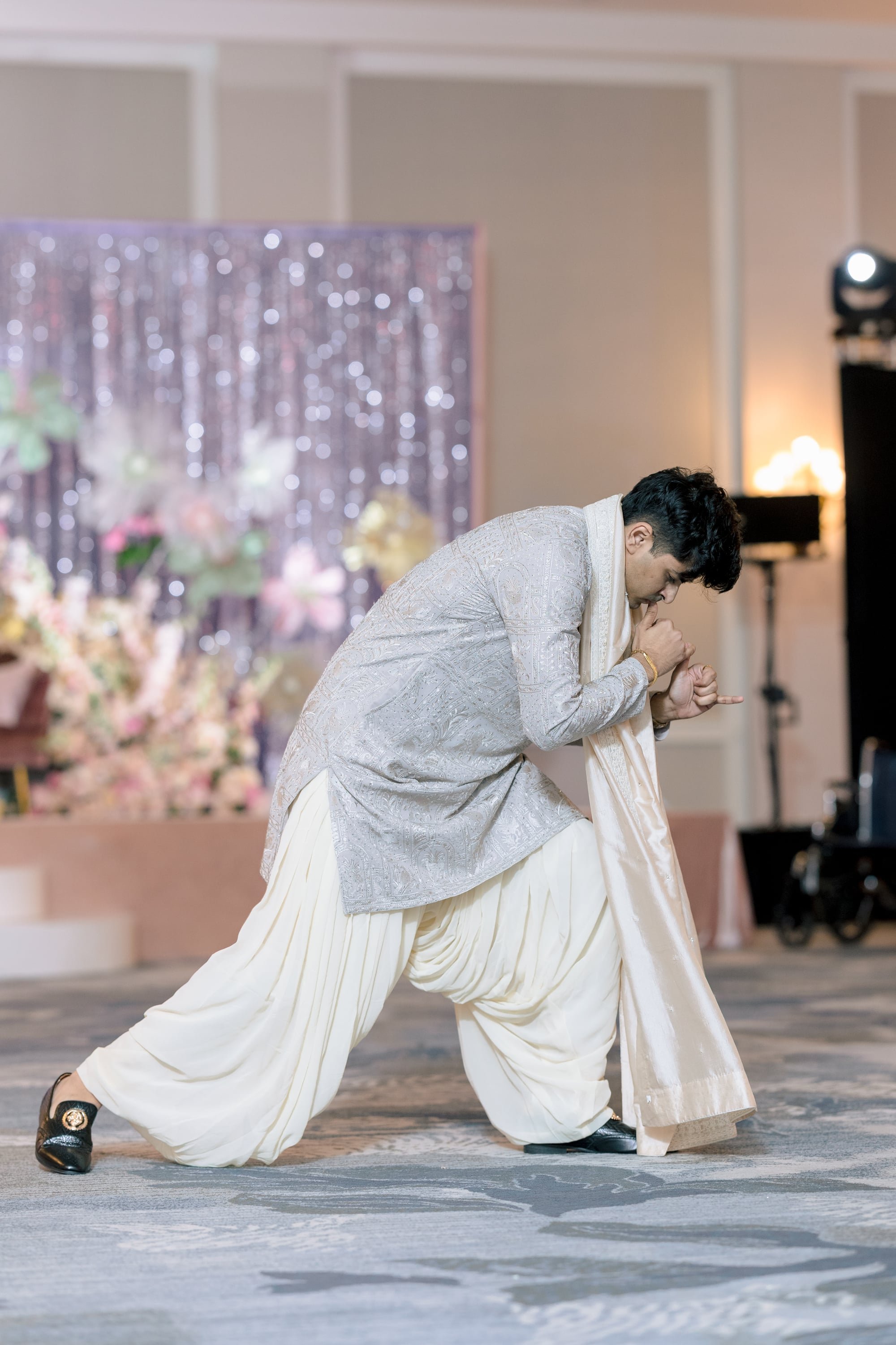 A man dressed in traditional Indian wedding attire is performing a dance move indoors. He is wearing a cream-colored sherwani with intricate embroidery and a matching dupatta. The background includes a curtain with sparkling decorations and floral ar