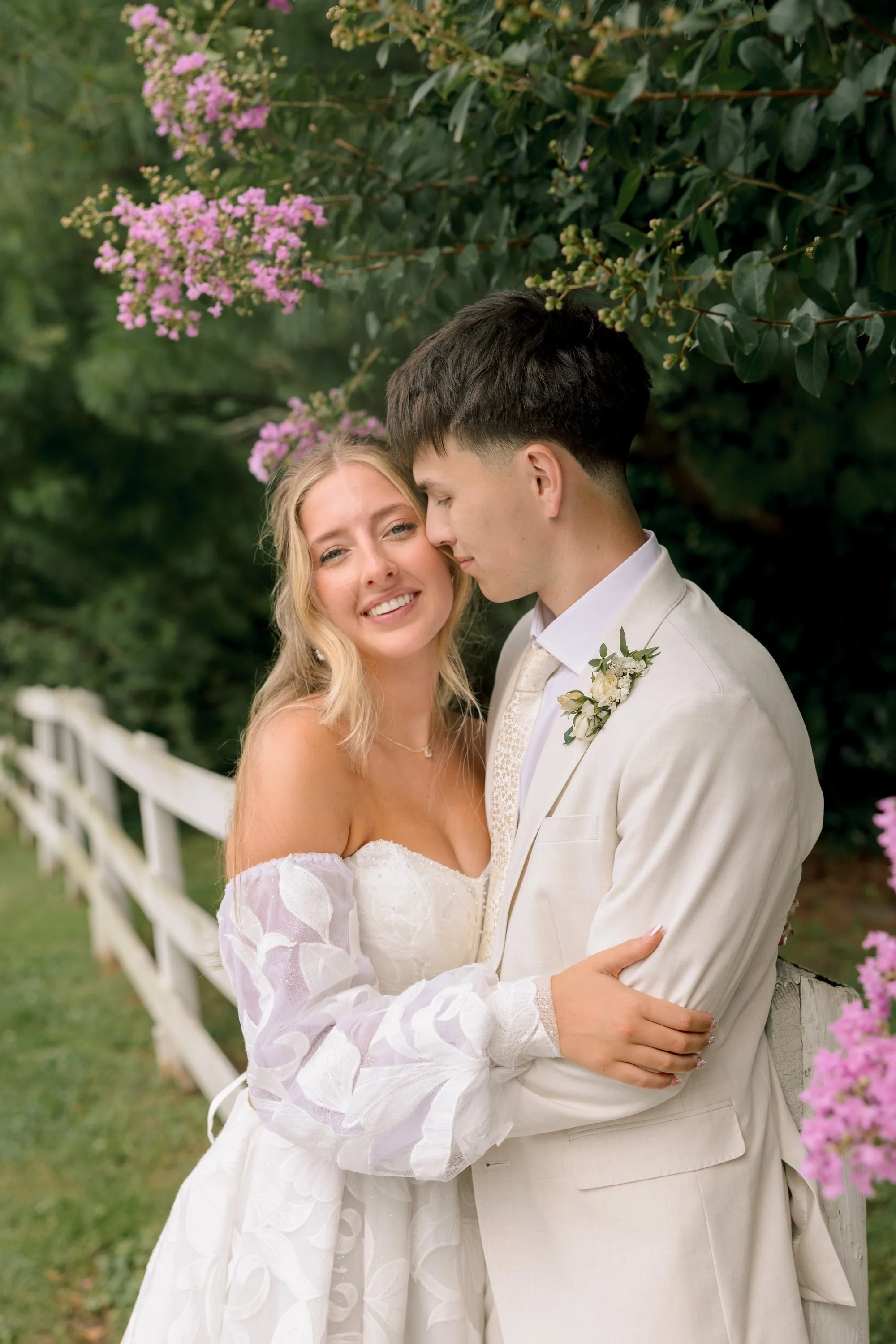 A bride and groom embrace each other outdoors near a white fence with pink flowering bush in the background.