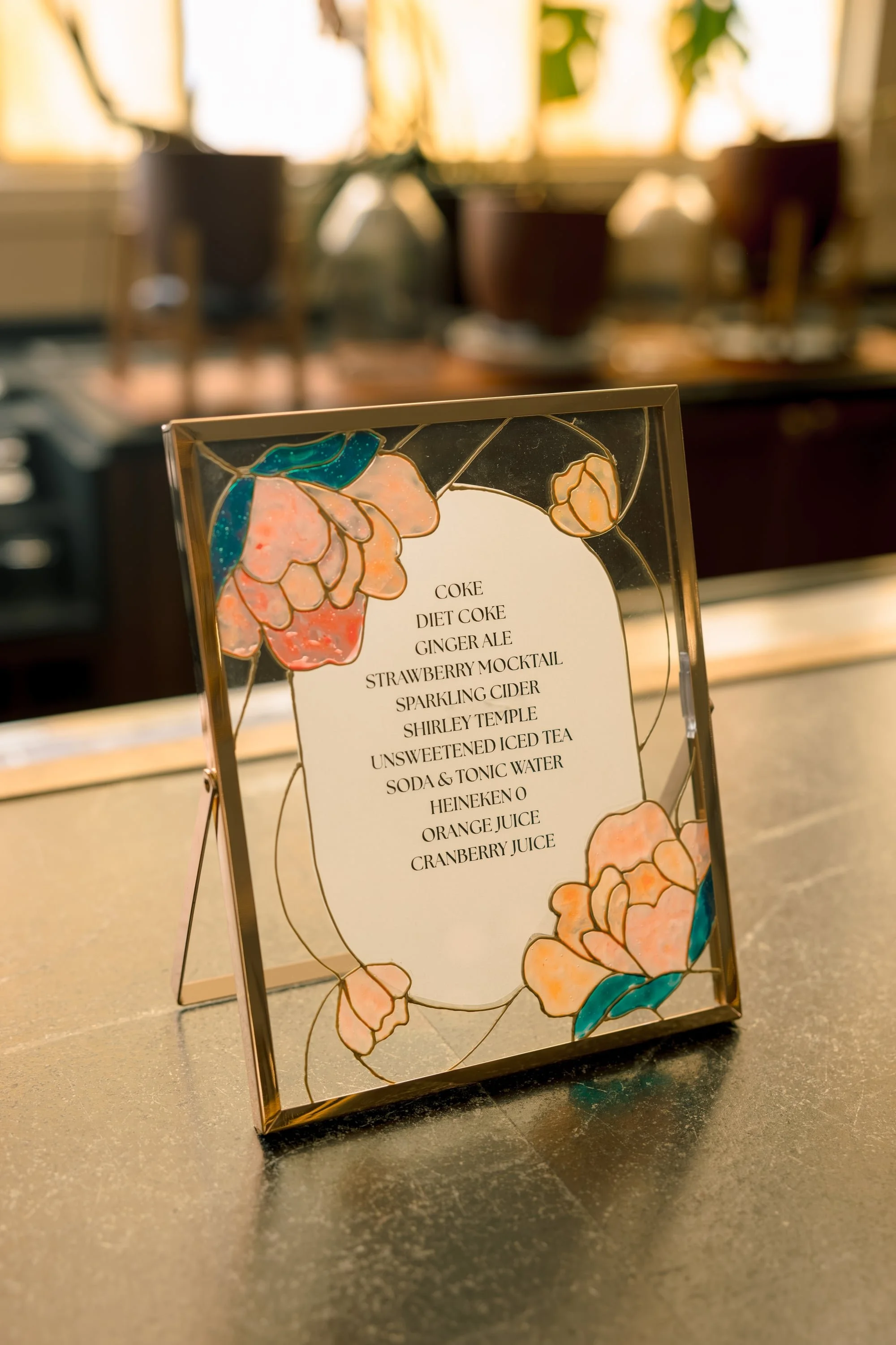 A decorative menu sign with a floral design on the border, listing drinks including Coke, Diet Coke, Ginger Ale, Strawberry Mocktail, Sparkling Cider, Shirley Temple, Unsweetened Iced Tea, Soda & Tonic Water, Heineken, Orange Juice, and Cranberry Jui