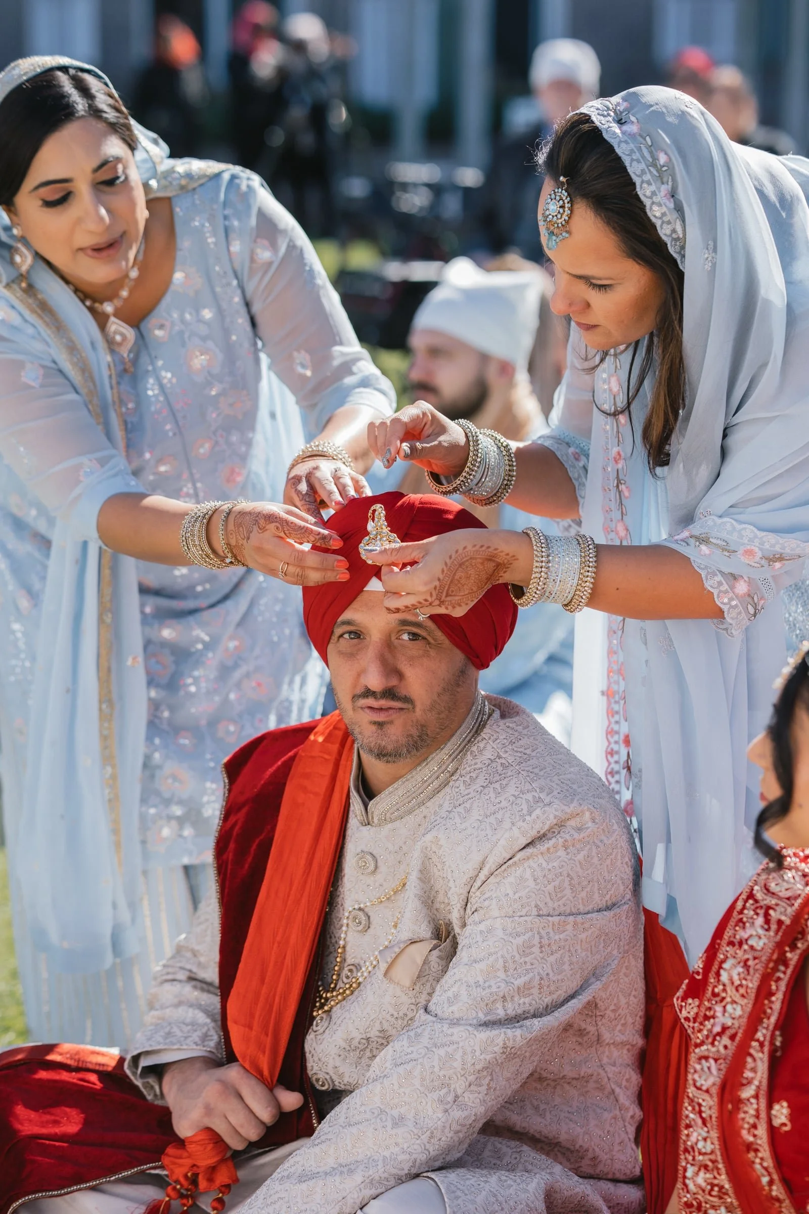 Two women in traditional South Asian attire performing a ritual or ceremony involving a man.