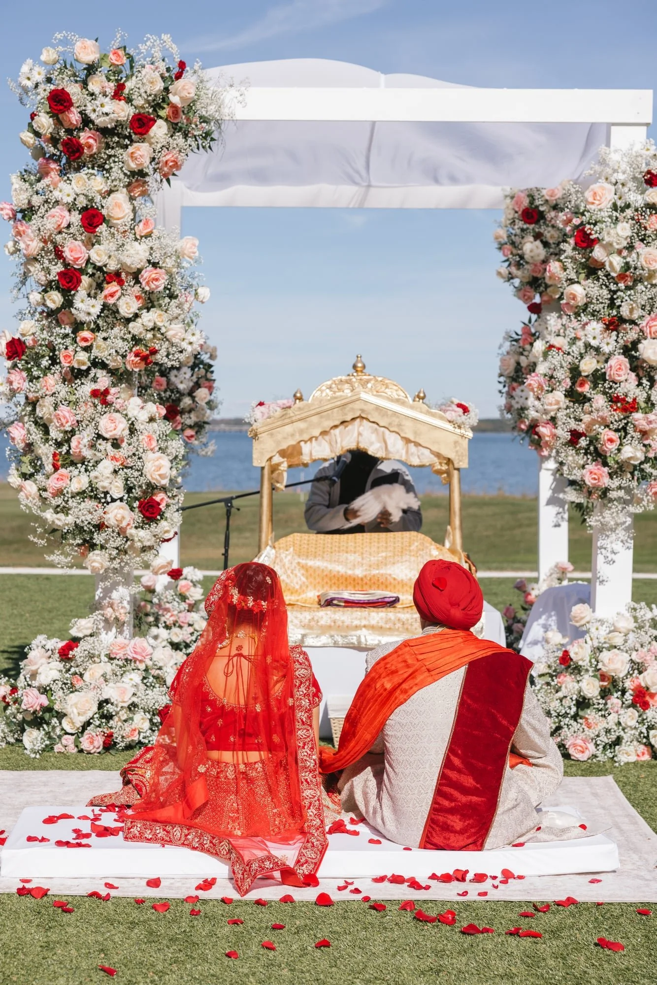 A traditional Indian wedding ceremony setup outdoors on a grassy area near water, featuring a floral arch with pink and white roses and white baby's breath, with a groom and a woman in red traditional attire seated on a white platform covered with ro