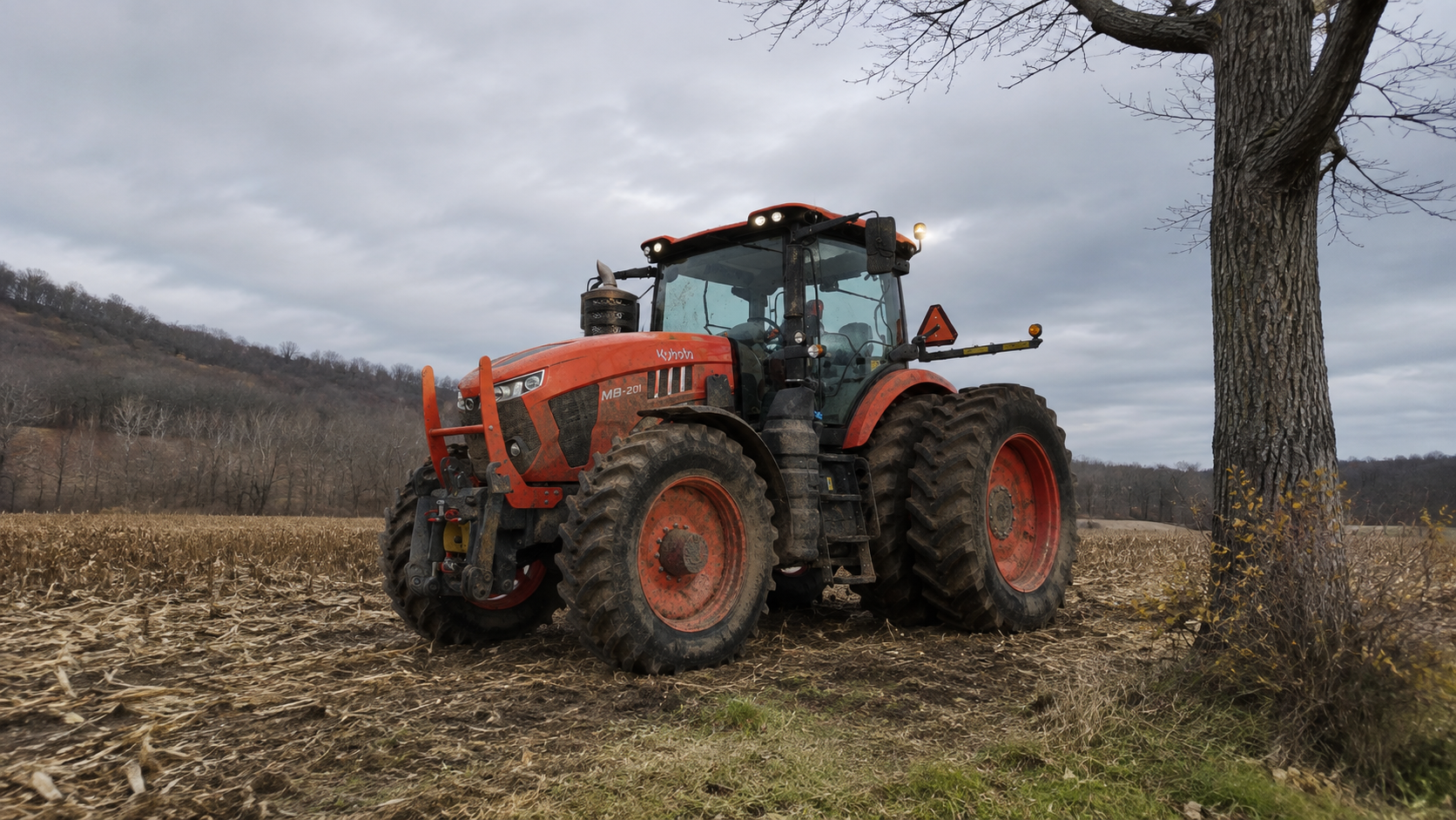 A red tractor parked near a leafless tree in a field with hills in the background on an overcast day.