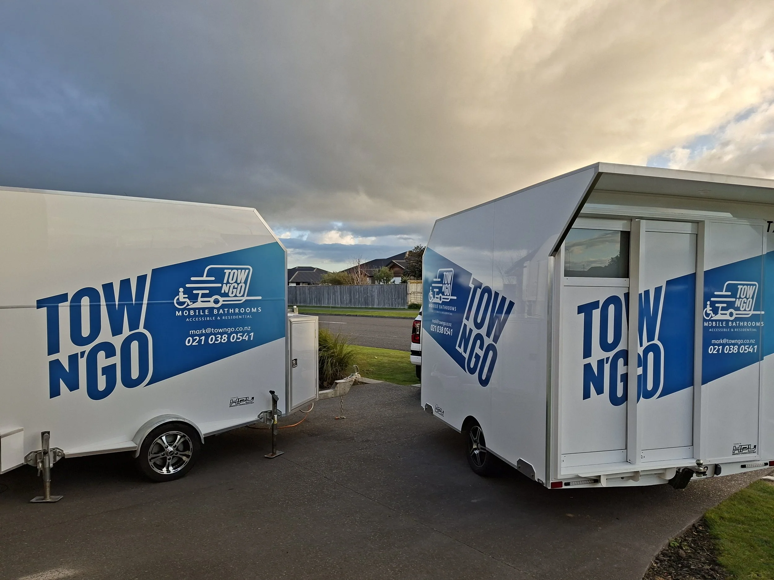 Two mobile bathroom trailers with blue and white branding parked outside on a cloudy day, with a house and trees in the background.