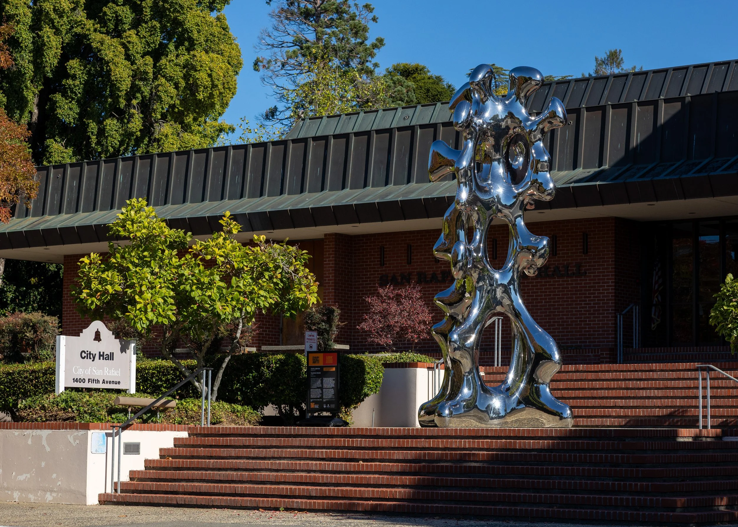 A shiny, reflective abstract sculpture in front of a city hall building with trees and a clear blue sky in the background.