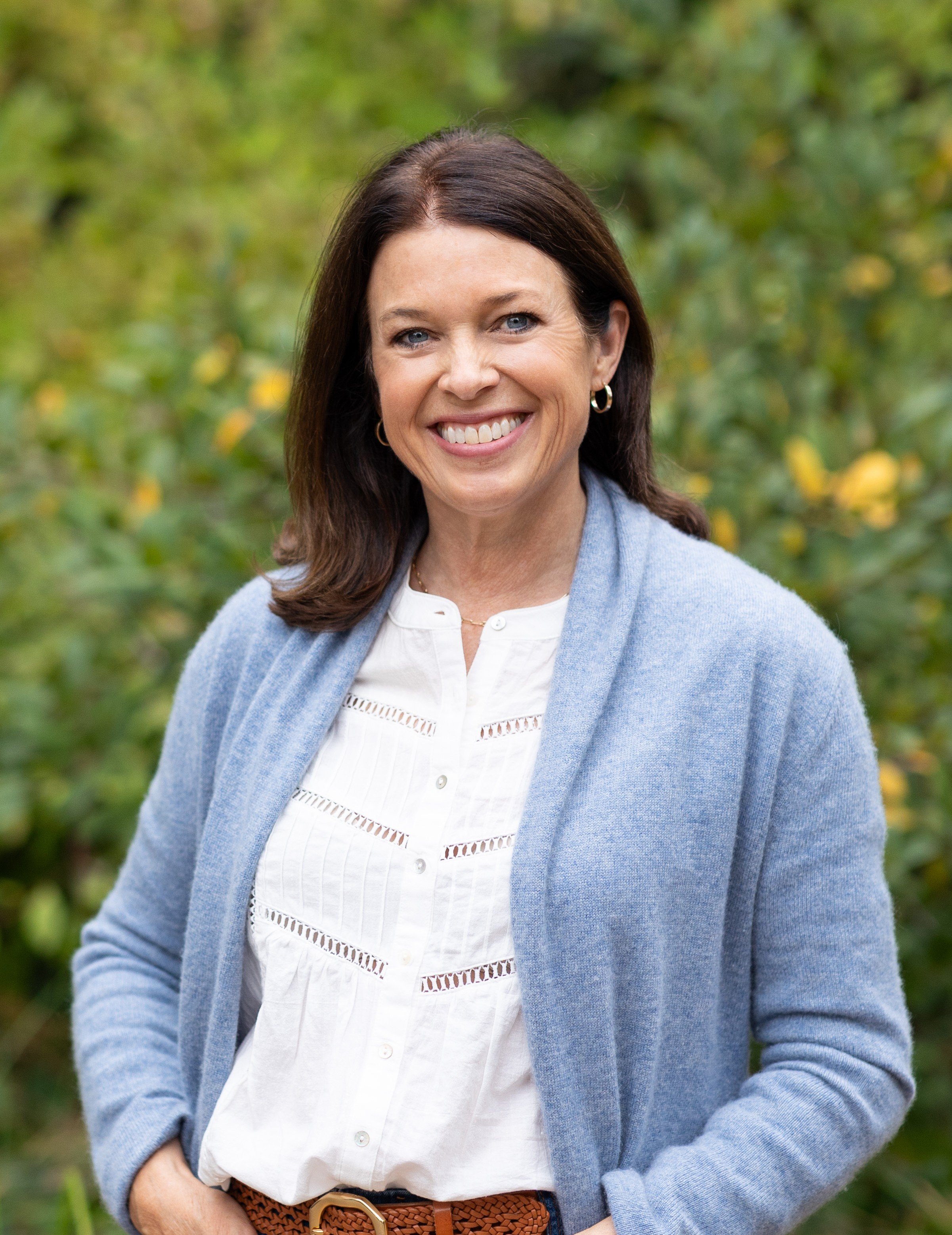 Close-up of a woman smiling outdoors, wearing a white shirt, a light blue cardigan, and a brown belt, with green foliage in the background.
