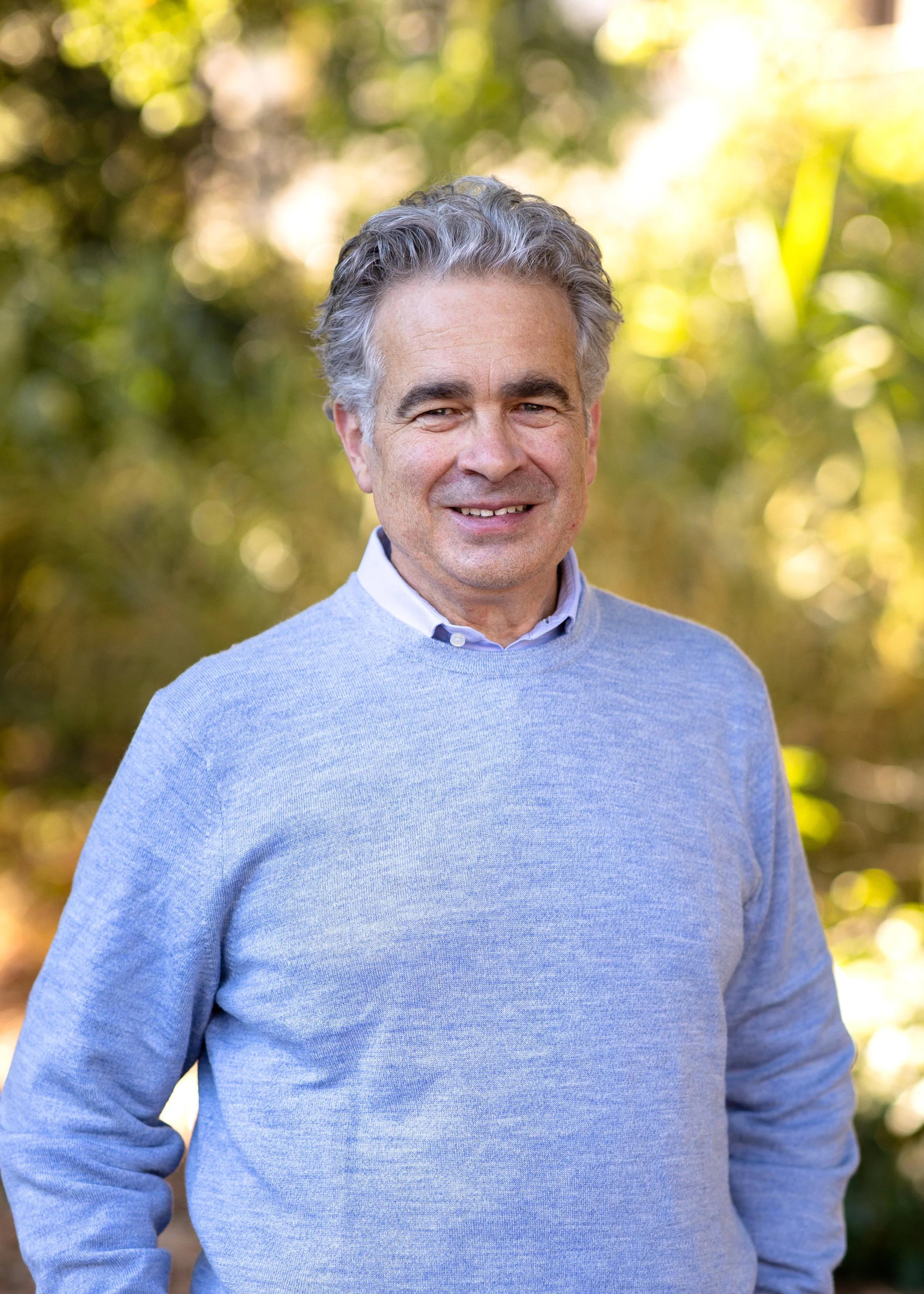 Older man with gray hair smiling outdoors, wearing a light blue sweater and collared shirt, with blurred green foliage background.