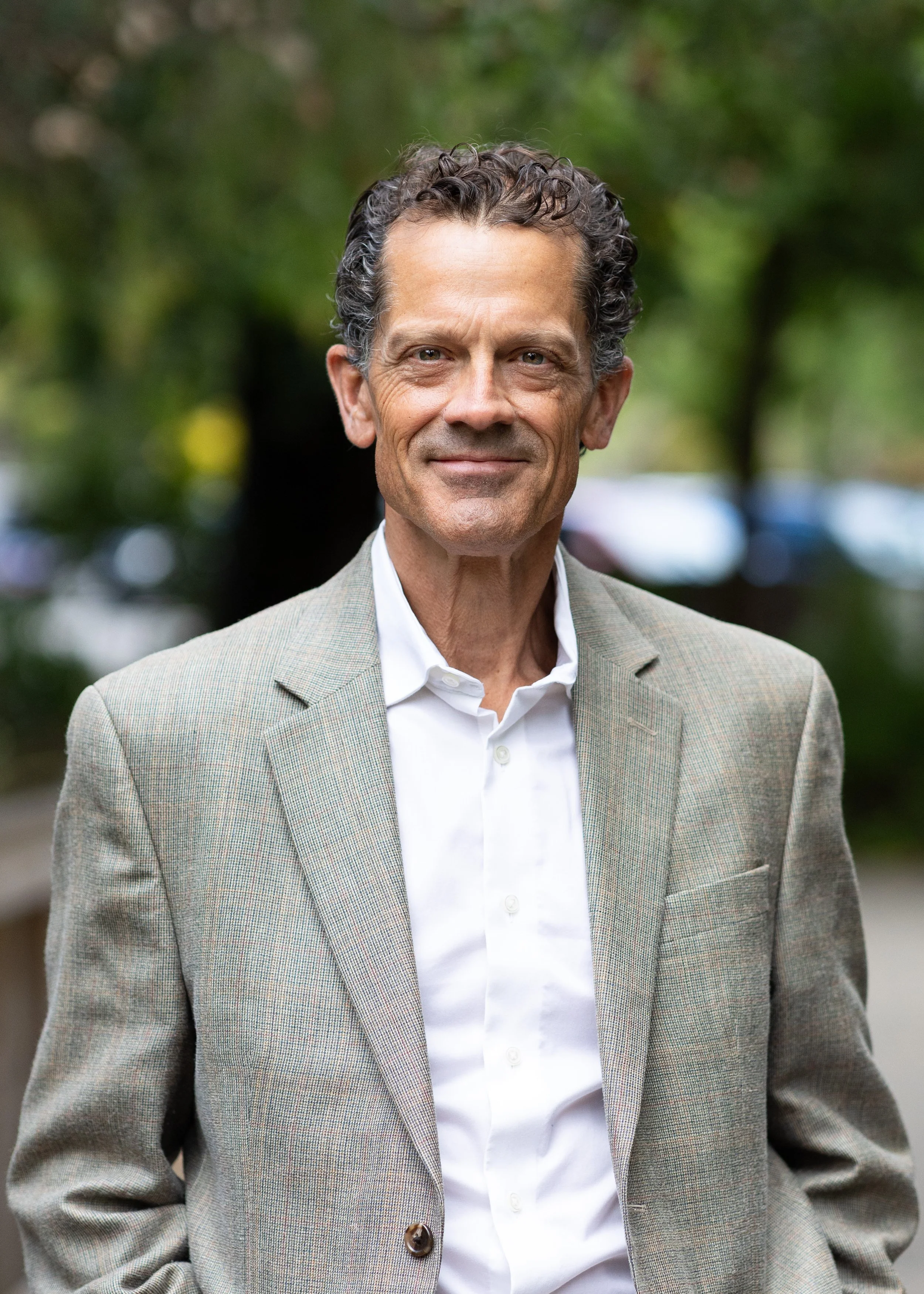 A middle-aged man with curly salt-and-pepper hair, wearing a light gray checkered blazer over a white button-up shirt, standing outdoors with a blurred background of green trees.