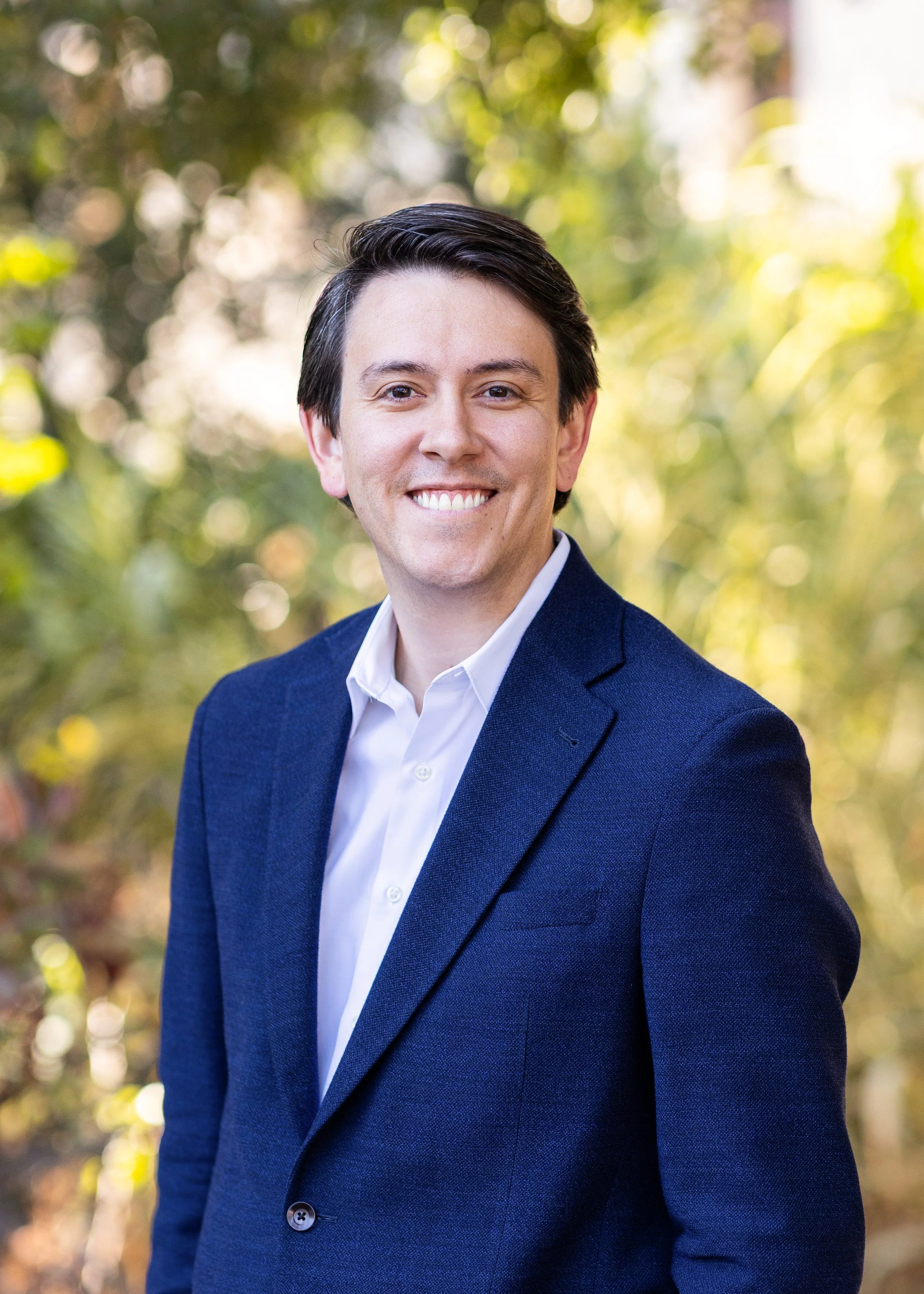 A man in a navy blue blazer and white shirt smiling outdoors with a blurred background of trees and sunlight.