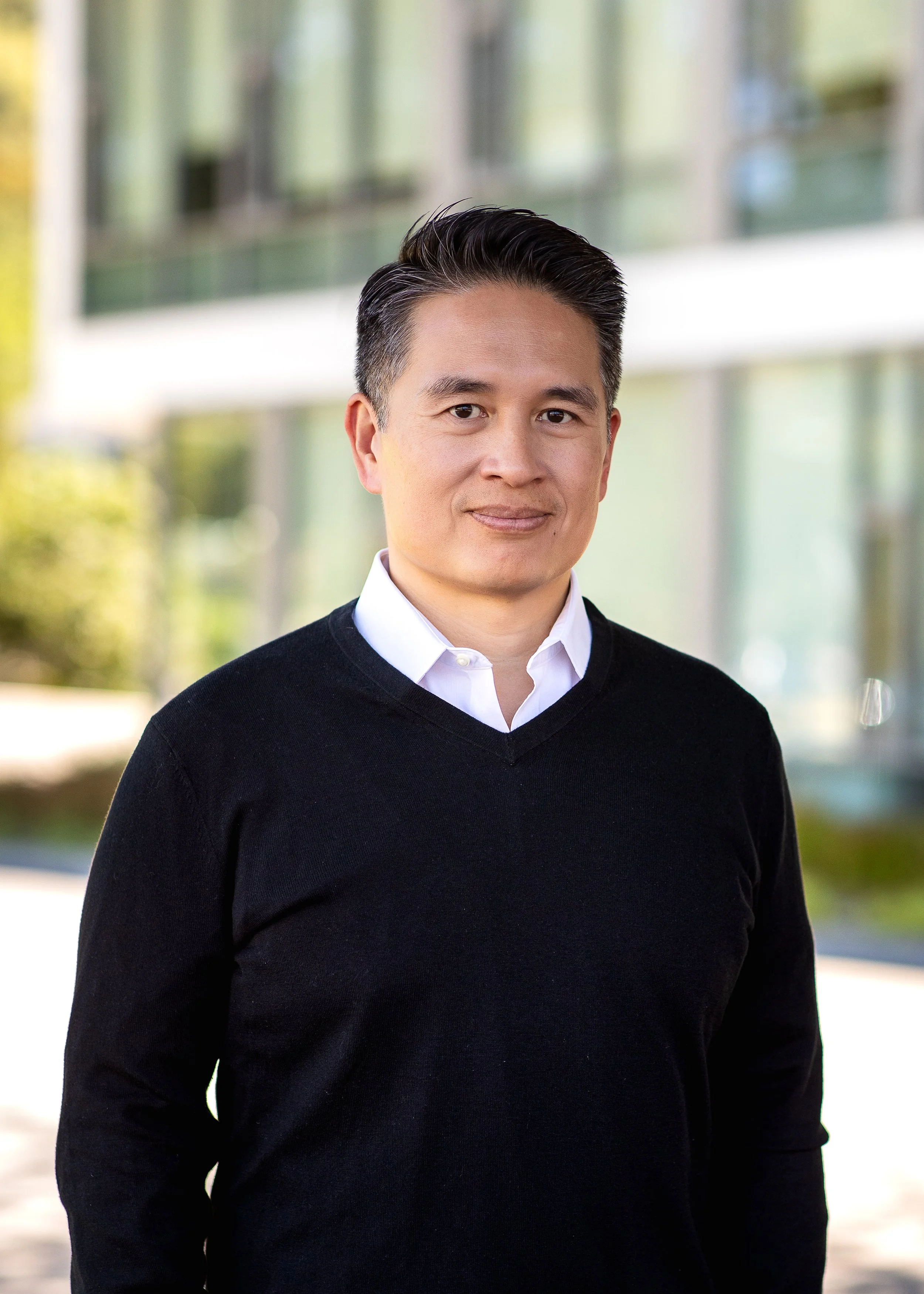 A man with short black hair, wearing a white collared shirt and black sweater, standing outdoors with blurred building and greenery in the background.