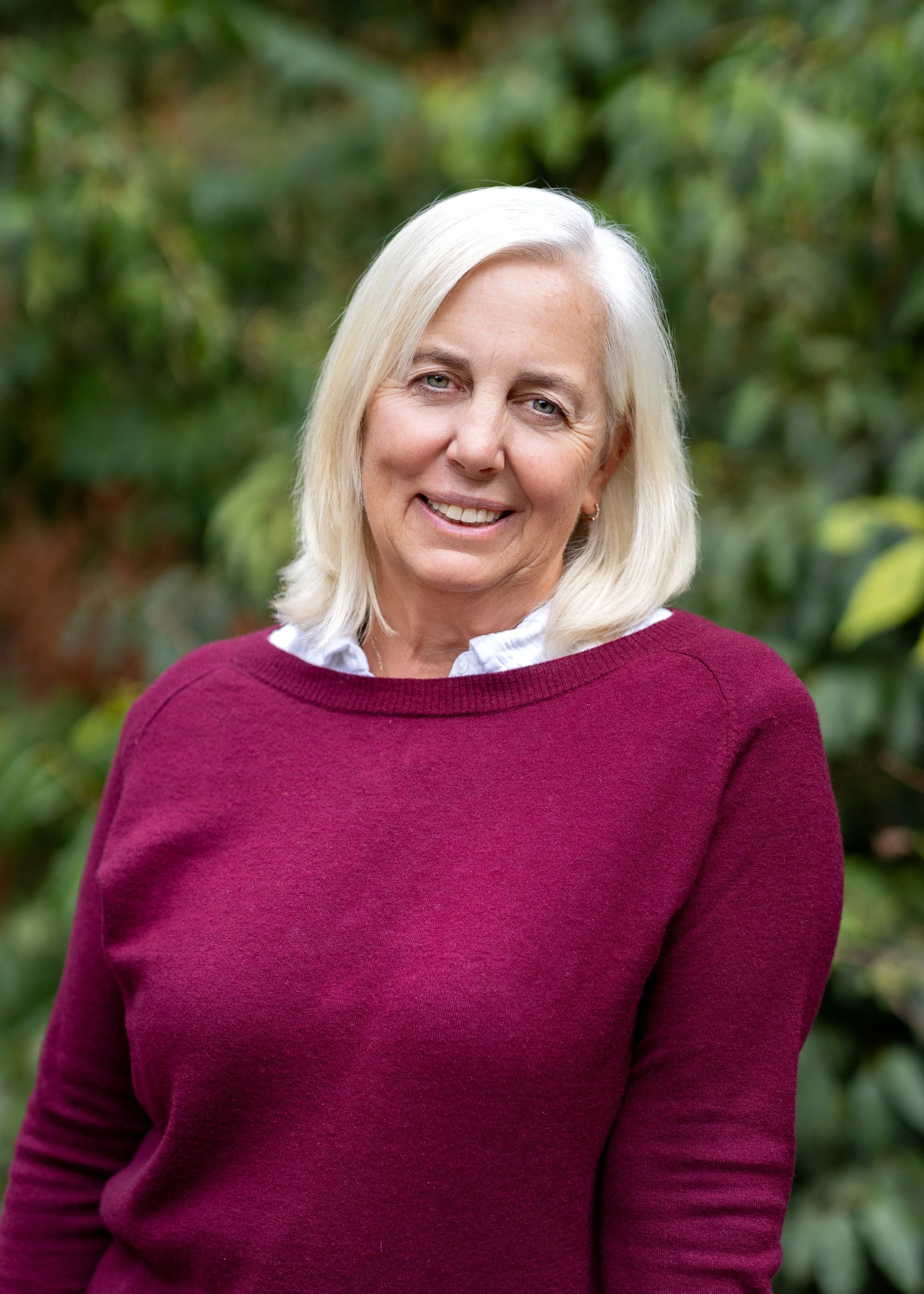 A smiling middle-aged woman with pale blonde hair, wearing a white collared shirt under a maroon sweater, stands outdoors with a blurred green foliage background.