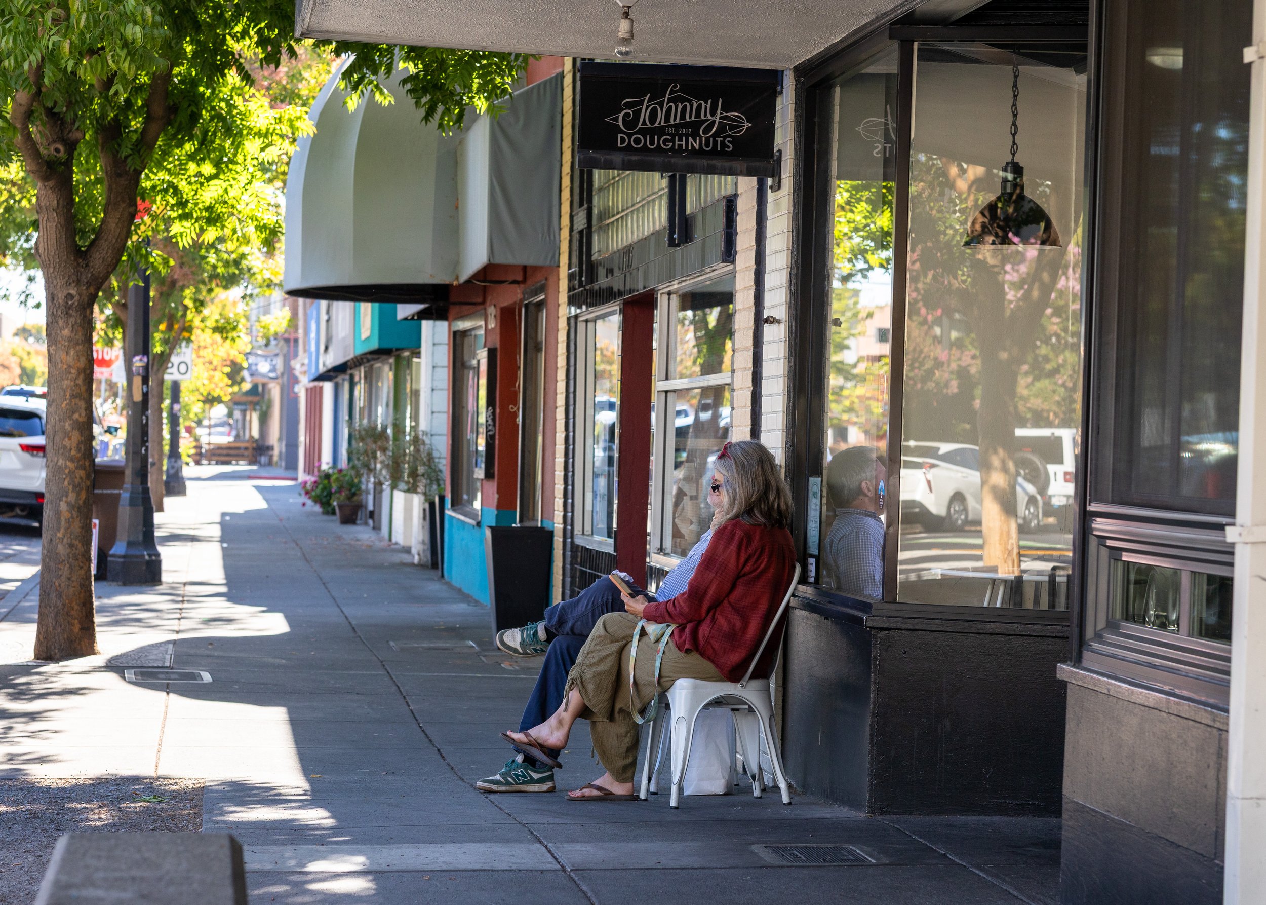A sidewalk scene with a woman sitting on a white bench reading something, and another person sitting next to her. They are in front of a storefront with large glass windows, a sign that reads 'Johnny Doughnuts', and trees lining the street.''
