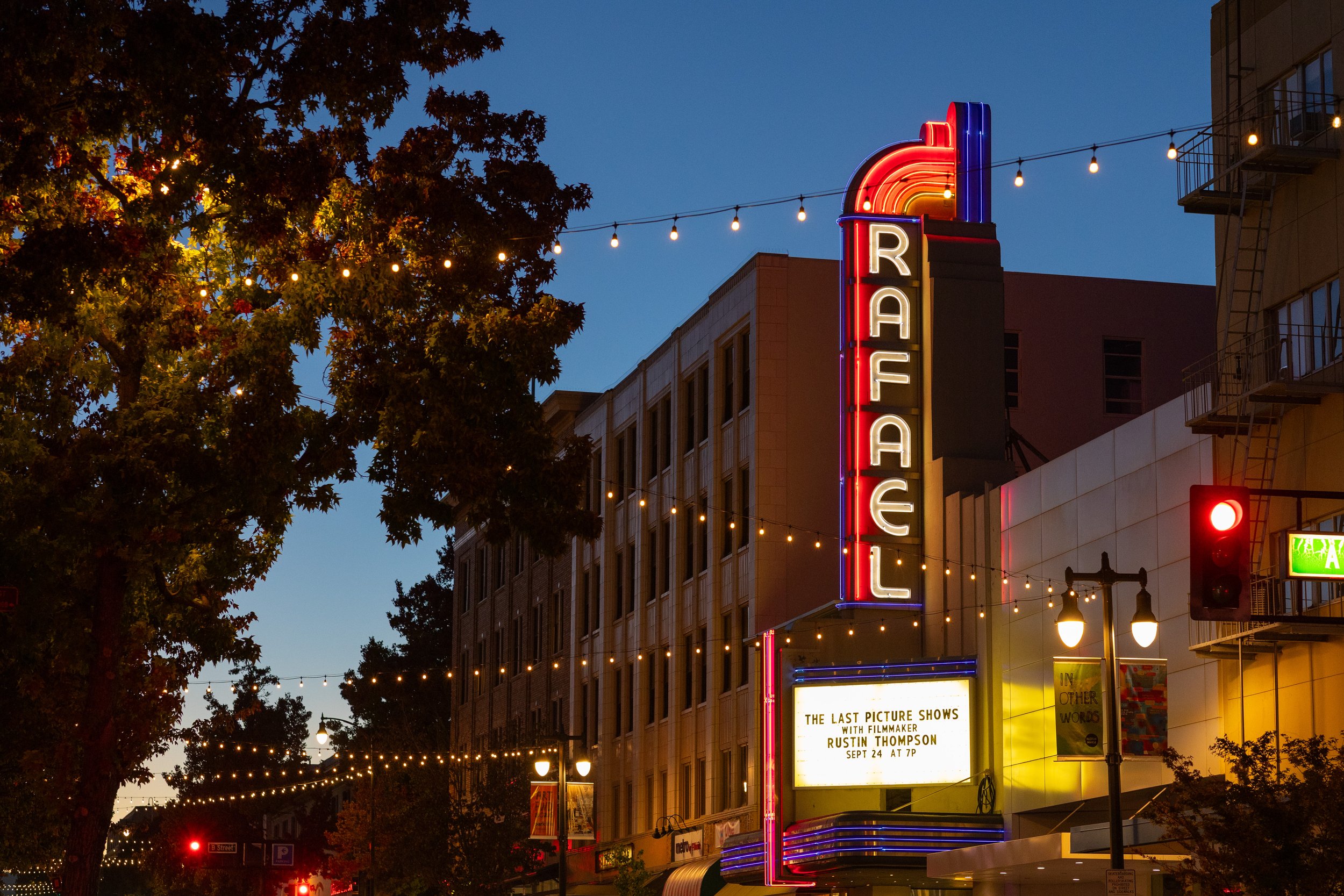 The image shows a city street at dusk with a tall neon sign that reads 'RAFAEL'. Downtown San Rafael, CA. String lights are hanging across the street, illuminating the area. There are trees with leaves visible and a building with windows and fire esc